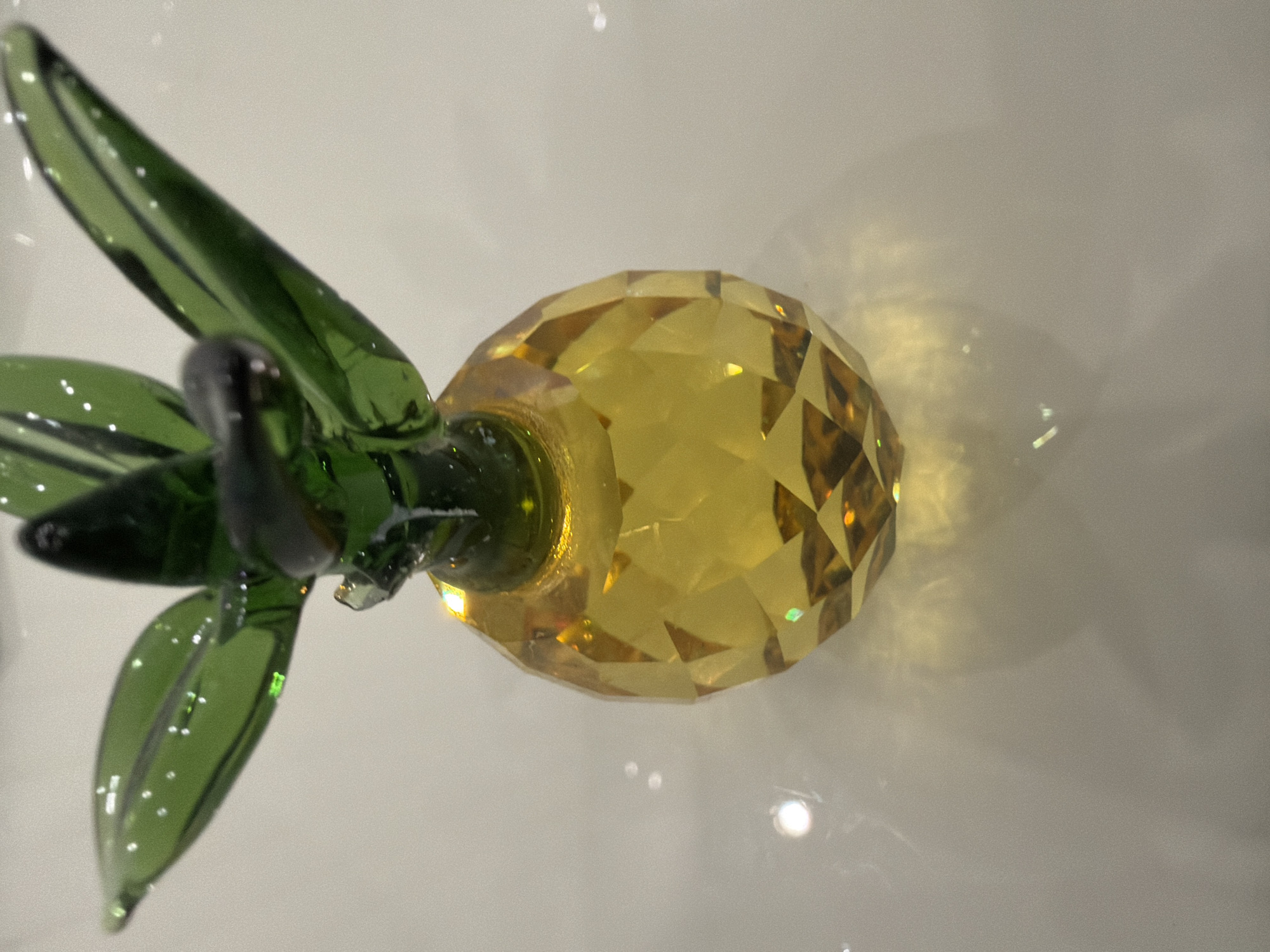A close-up, eye-level shot shows a clear yellow glass pineapple figurine on a white, reflective surface. The pineapple has a faceted body and green glass leaves at the top. The surface reflects the light and the figurine, creating a soft shadow beneath it. There is no indication of the city of Podgumer, Bulgaria in the image.