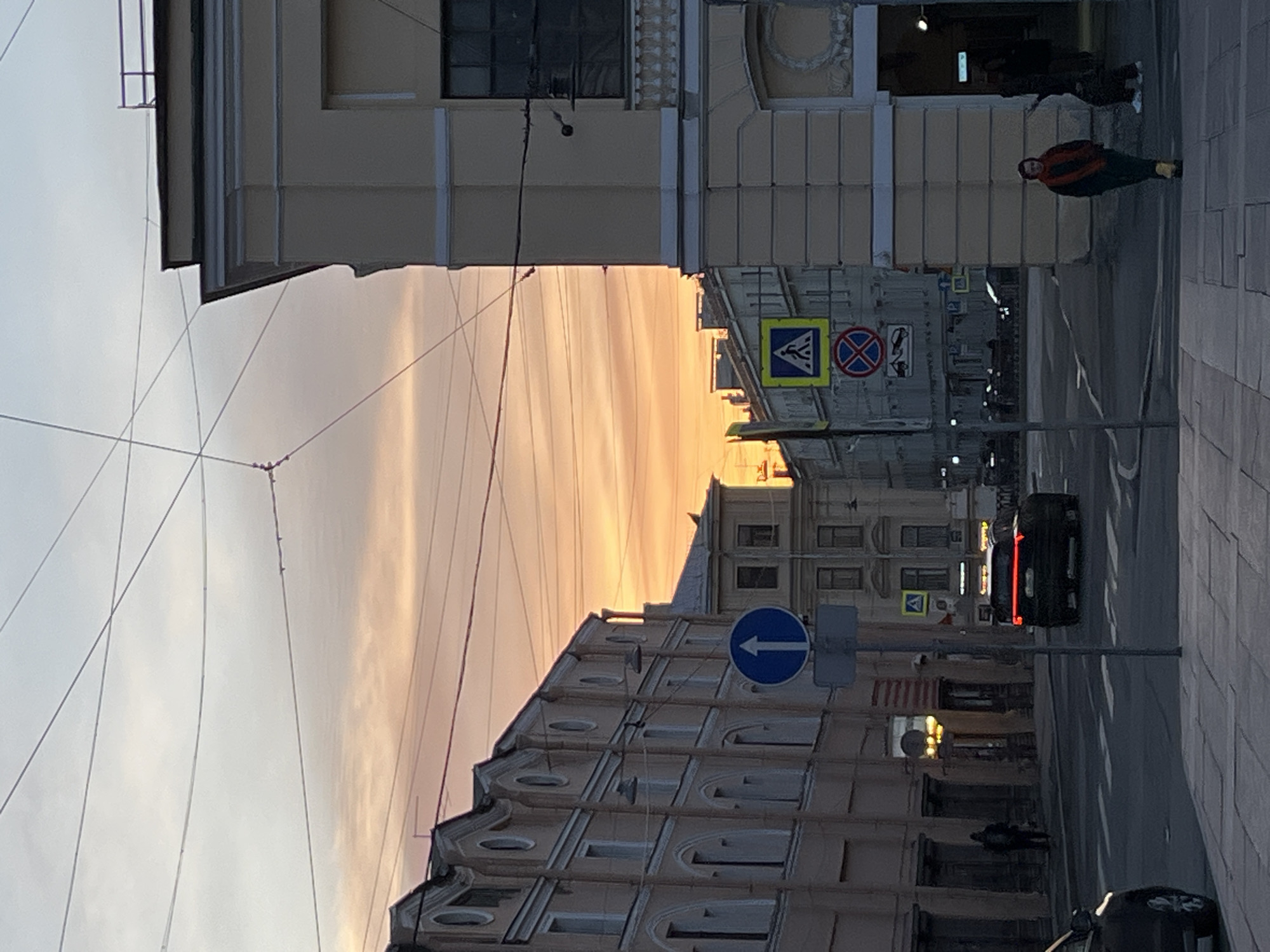 An urban street scene unfolds during dusk or dawn, characterized by a soft, warm golden light from a low sun reflecting on the clouds and sky. The sky transitions from a light, hazy grey at the top to a vibrant, streaky golden-orange towards the horizon. Numerous thin overhead wires crisscross the sky, suggesting trolleybus lines or utility cables.
The street is flanked by multi-story buildings of classical architectural style, featuring decorative elements like columns, cornices, and arched windows, particularly noticeable on the building in the foreground on the left. The buildings are lit unevenly, with some parts in shadow and others catching the warm light.
In the lower half of the image, a dark asphalt road is visible, marked with white lines. Paved sidewalks run along both sides of the street. On the right-hand sidewalk, a person is walking, wearing a distinctive orange or red jacket, dark pants, and yellow shoes. Further down the street, on the left sidewalk, another blurry figure is discernible.
Several traffic signs are present: on the left side of the street, a yellow square sign with a black triangle and a walking person symbol indicates a pedestrian crossing ahead. Adjacent to it is a red circular sign with a blue 'X', signifying 'No Stopping'. Closer to the foreground, a blue circular sign with a white arrow pointing to the left is mounted on a pole, indicating a mandatory turn. A black car with illuminated red taillights is seen on the road, suggesting low light conditions and active traffic. No explicit text is readable on any signs or buildings.