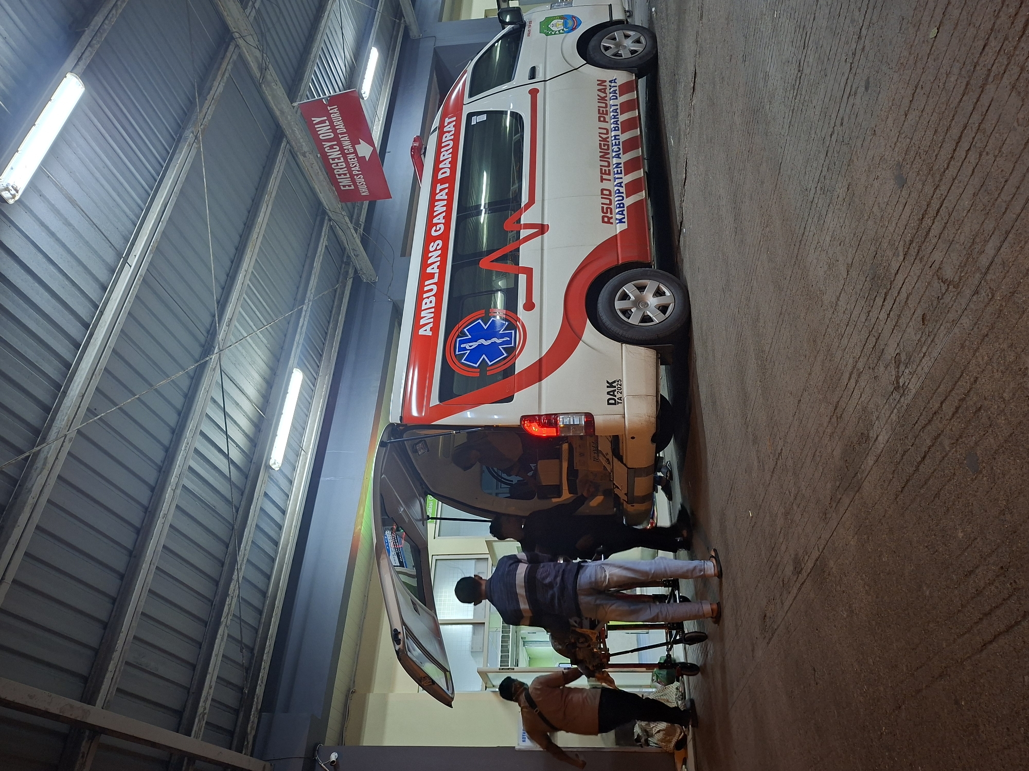 The image shows an ambulance outside of an emergency room in Banda Aceh, Indonesia. The ambulance is white with red striping and lettering in Indonesian. Two men are visible near the open rear door of the ambulance, presumably tending to a patient on a gurney. Above the ambulance, a sign in Indonesian and English reads "EMERGENCY ONLY / KHUSUS PASIEN GAWAT DARURAT" (Emergency Only / For Emergency Patients Only). The scene is set in a covered loading area with a metal corrugated roof and fluorescent lighting. The hospital is identified on the ambulance as RSUD Teungku Peukan, Kabupaten Aceh Barat Daya (Teungku Peukan Regional General Hospital, West Aceh Southwest Regency).