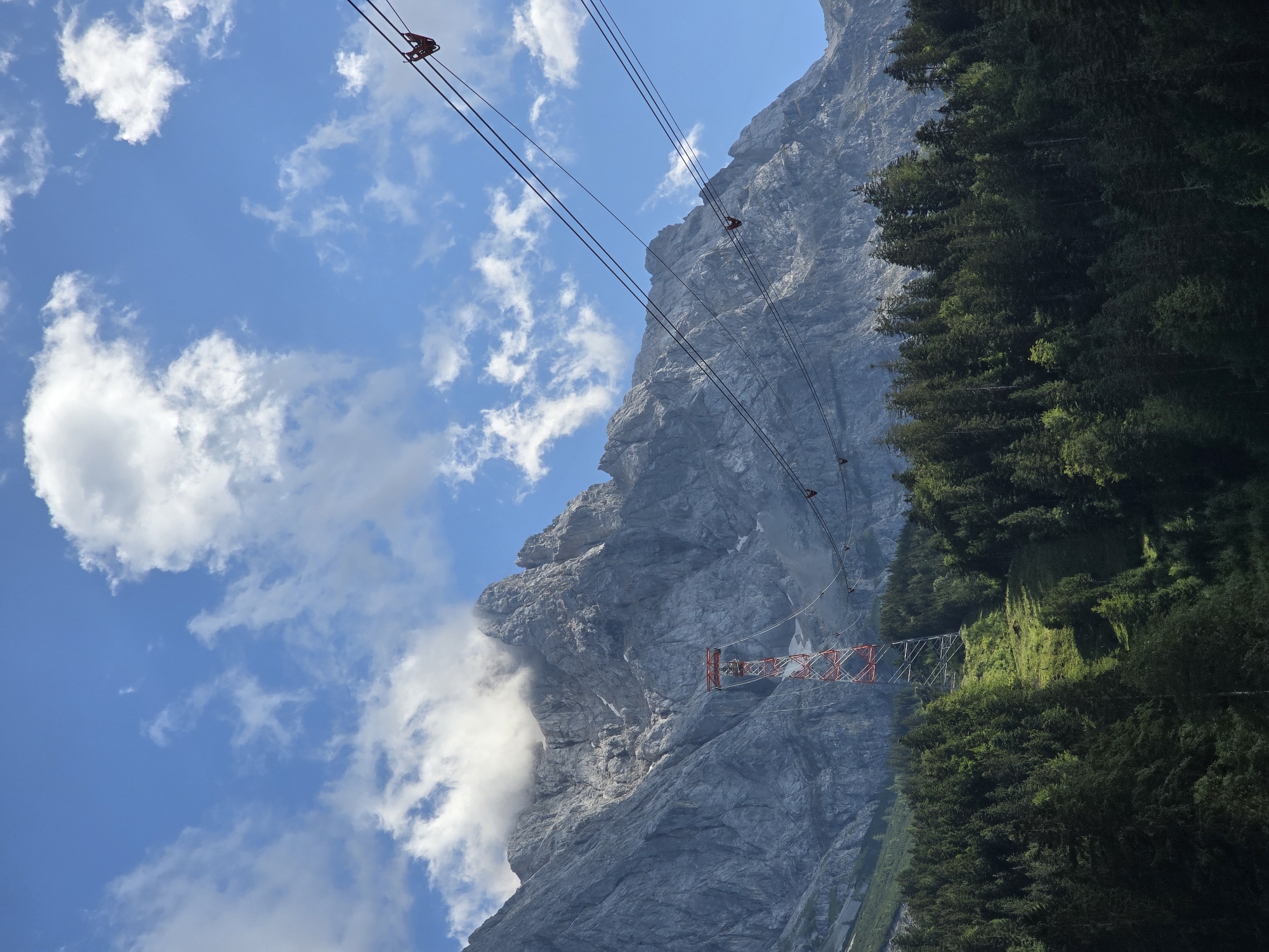 The image depicts a dramatic mountain landscape on a sunny day with scattered clouds. A prominent element is a cable car system, with thick cables stretching across the frame and two red cable car cabins visible. One cabin is closer to the foreground, suspended from the cables, while another is further up the mountain. A tall, red and white pylon supports the cables on the mountainside.
The mountain itself is a massive, gray, rocky peak, with steep, imposing cliffs. Patches of snow are visible on the higher elevations of the mountain. A dense forest of dark green evergreen trees covers the lower slopes and a portion of the mountainside to the right of the image. Wisps of white clouds are clinging to the mountain face, adding a sense of height and atmosphere. The sky is a vibrant blue, indicative of fair weather.
The scene suggests an outdoor activity, likely tourism or recreation, involving the cable car. The time of day appears to be daytime, given the bright sunlight casting shadows and illuminating the landscape. The location is identified as Grainau, Germany, a region known for its alpine scenery. There is no visible text in the image. The overall impression is one of majestic natural beauty combined with human engineering, offering a sense of adventure and awe.