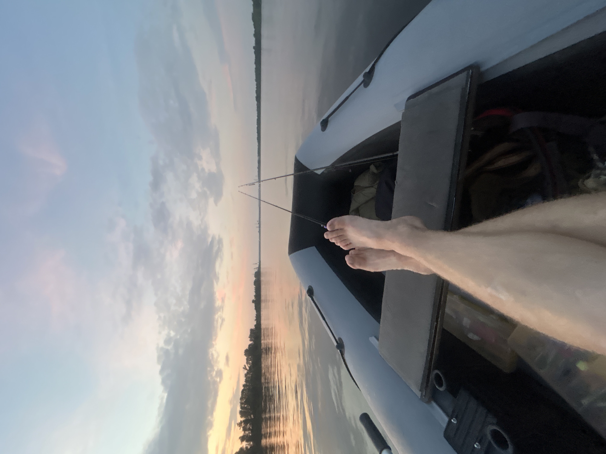 In the city of Noviy, Russia, at dusk, a first-person perspective shot shows a person's bare legs and feet resting on the floor of a small, grey inflatable boat. The boat is on a wide, calm river, reflecting the soft, pastel colors of the twilight sky. Two fishing rods are propped up in the boat. The shoreline, lined with trees, is visible in the distance, with industrial structures faintly appearing on the horizon. The sky transitions from a pale orange and pink near the horizon to a soft blue with scattered clouds.