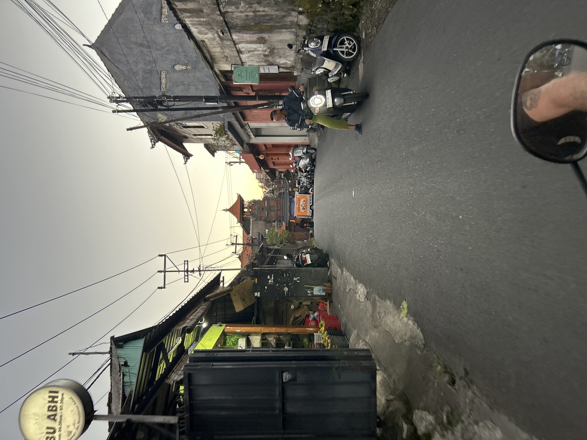 The image shows a street scene in Ubud District, Indonesia. A man is riding a scooter down a narrow street lined with buildings. The buildings appear to be shops or residences, with some of them having open storefronts displaying goods. There are several other scooters parked on the side of the street. In the distance, a traditional Balinese gate is visible. The sky is overcast, suggesting it might be early morning or late evening. The overall impression is of a busy, local street in a Southeast Asian city.