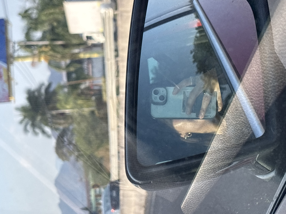 This image, captured from inside a vehicle, presents a dual perspective: the exterior environment through the windshield and a reflection in the passenger-side mirror. The location is identified as Oaxtepec, Mexico.
The exterior view through the windshield depicts a sunny daytime scene. In the background, prominent mountains are visible on the left. The mid-ground features a multi-lane road or highway separated by a concrete median barrier. Dense green vegetation, including multiple palm trees, lines the roadside. A dark-colored sedan is partially visible on the left, moving away from the viewer. On the right, a large billboard with red and blue elements is mounted on a yellow pole, and a white delivery truck is partially visible beyond the barrier.
The reflection in the side mirror, which occupies the lower half of the image, displays the left profile of an individual. This individual has dark hair and wears a small, circular gold-colored hoop earring on their visible ear. Their right hand is holding a light-colored smartphone vertically, revealing a dual-lens camera system on the upper left rear of the device. The hand wears multiple rings on different fingers. A textured, dark-colored surface, consistent with a vehicle's dashboard or door panel, is visible in the lower left of the reflection, and the vehicle's exterior color, a dark metallic red or maroon, is reflected in the mirror's frame.