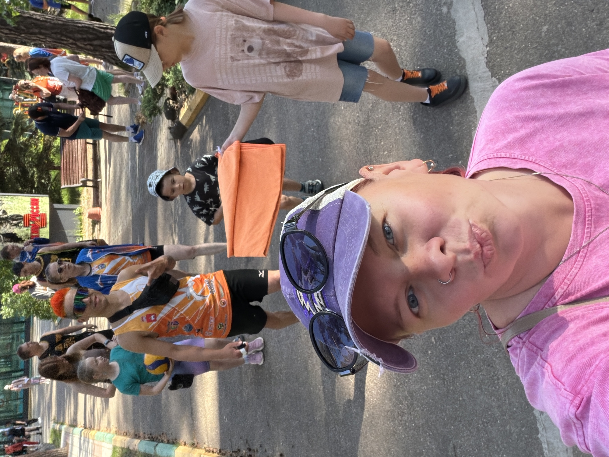 The image shows a group of people walking on a paved path in Chaykovsky, Russia. The camera appears to be held by a person wearing a pink shirt and a purple baseball cap with sunglasses perched on the brim. This person is looking into the camera, making a kissing face. Behind them, a group of people are walking in the same direction. There are children and adults visible. One child is wearing a pink shirt with a bear graphic and a black and white baseball cap, while another is wearing a dinosaur-print shirt and carrying an orange towel. Some of the adults are wearing athletic wear with the word "VAKAR" on their shirts. One person is holding a volleyball. Trees and green foliage are visible on the side of the path, indicating a park-like or recreational area. A line of buildings is partially visible in the background. The general impression is one of a casual group outing.
