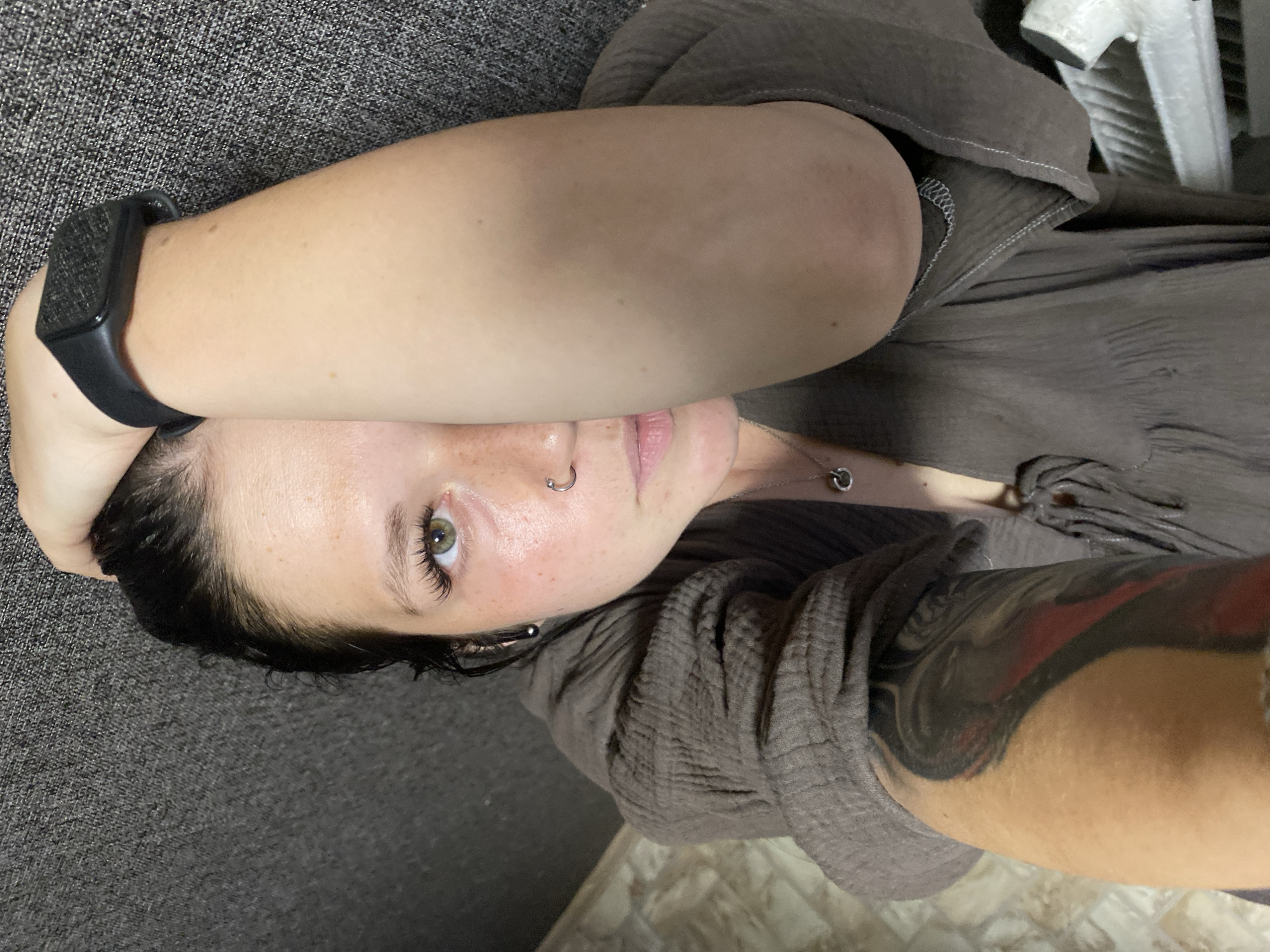 An overhead medium shot shows a woman lying down with her left arm extended, covering part of her face. Her right arm is bent and resting on her head. She has dark hair pulled back and is wearing a brown gauzy top with a silver necklace. She has a nose ring and earrings. A black smartwatch is visible on her right wrist. Her left arm has a large tattoo with red and black elements. The background consists of a grey textured surface and a tiled floor. The location is specified as Chaikovsky, Russia.