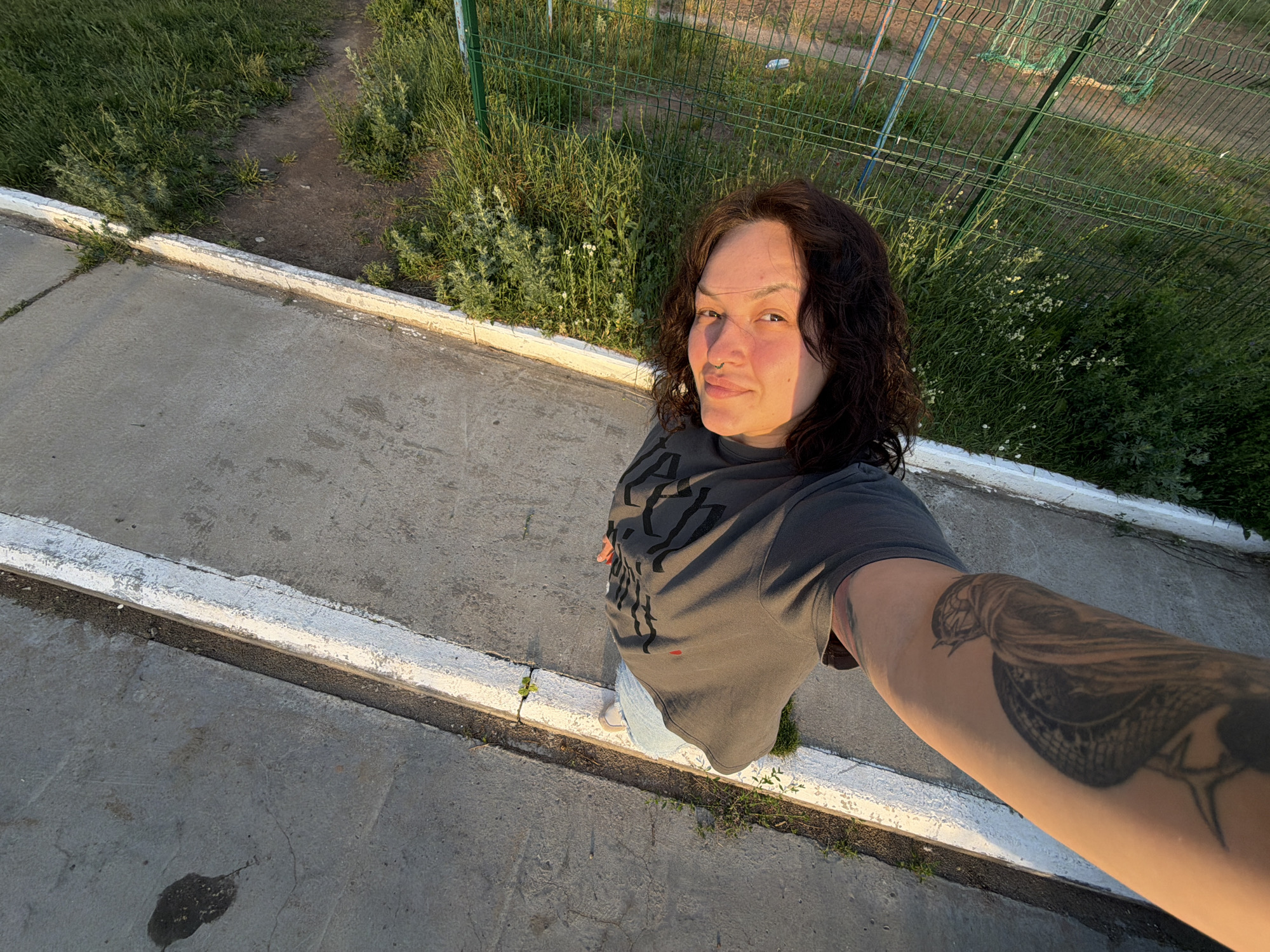 The image shows a person taking a selfie, holding the camera with their left arm, which displays a tattoo of a bird on the forearm. The person is standing on a concrete surface next to a white-painted curb. They are wearing a dark gray t-shirt with a graphic design. Their face is illuminated by the warm light of the setting sun, casting a reddish glow on their skin. The background consists of green grass and a green metal fence, suggesting an outdoor location, possibly a park or sports field. The perspective is from above, looking down at the person and the surrounding area. The image is oriented with the person's head at the bottom, suggesting it was taken in portrait mode and rotated. Without additional information, the specific location in Chaikovsky, Russia cannot be determined, but the scene depicts a casual outdoor setting during golden hour.