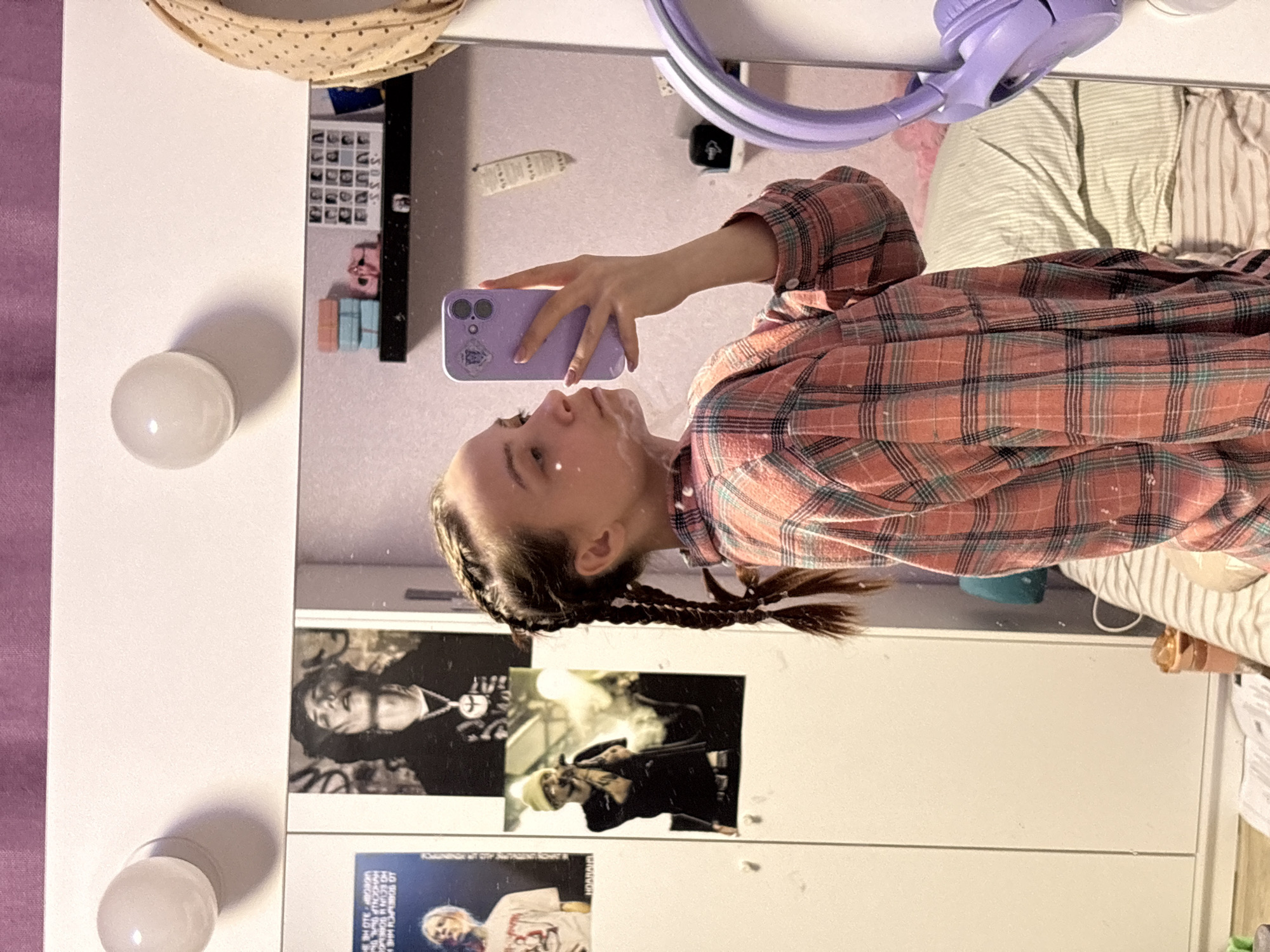 A young female is captured taking a mirror selfie with a purple smartphone in an indoor setting, likely a bedroom or personal space in Прикамский, Russia. The image is taken from the reflection in a white vanity mirror, which features several round, white light bulbs on its frame.
The central subject is a young woman, visible from the chest up. She has light skin and dark hair styled in two long, tightly woven braids. She wears a long-sleeved, plaid shirt in shades of orange-red, black, and light blue-green. Her right hand, with light-colored nail polish, holds a purple smartphone in a clear case with a small sticker on the back. Her gaze is directed towards the phone screen, focusing on the selfie.
In the background reflection, behind the woman, various items are visible. A pair of lavender over-ear headphones rests near the top right. A striped pillow, light green and white, and crumpled bed sheets are seen, suggesting a bed is nearby. The mirror also reflects several posters or pictures taped to a surface. These include a grid of small black and white images, and larger black and white photos of individuals, possibly musicians. One poster shows a person with a necklace, another depicts a person in a beanie holding a microphone, and a third displays a blonde individual in a white t-shirt. Some small, light blue and pink boxes or containers are also reflected. There is faint, illegible text on some of the reflected posters. A beige fabric with polka dots is draped near the top left corner of the reflected scene. The overall atmosphere suggests a personal and casually decorated space.