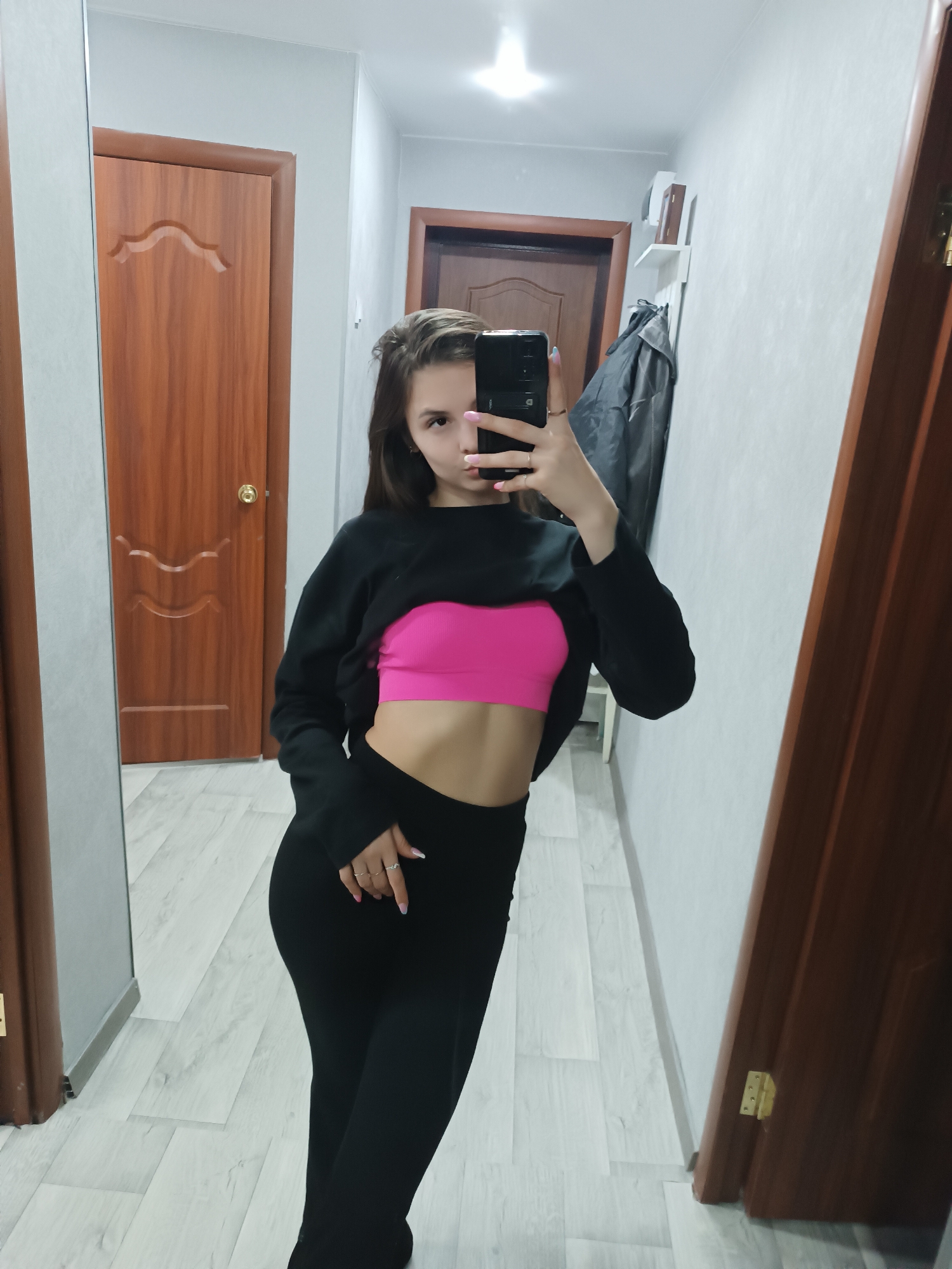 A young woman stands in a hallway, taking a mirror selfie. She is wearing black pants, a black cropped sweatshirt, and a bright pink sports bra. The hallway has light-colored walls and flooring. There are two closed wooden doors visible, one on either side of the hallway. A third door is partially visible at the end of the hallway. A coat hangs on a wall-mounted rack near the third door. Based on metadata, this image was taken in Chaikovsky, Russia.
