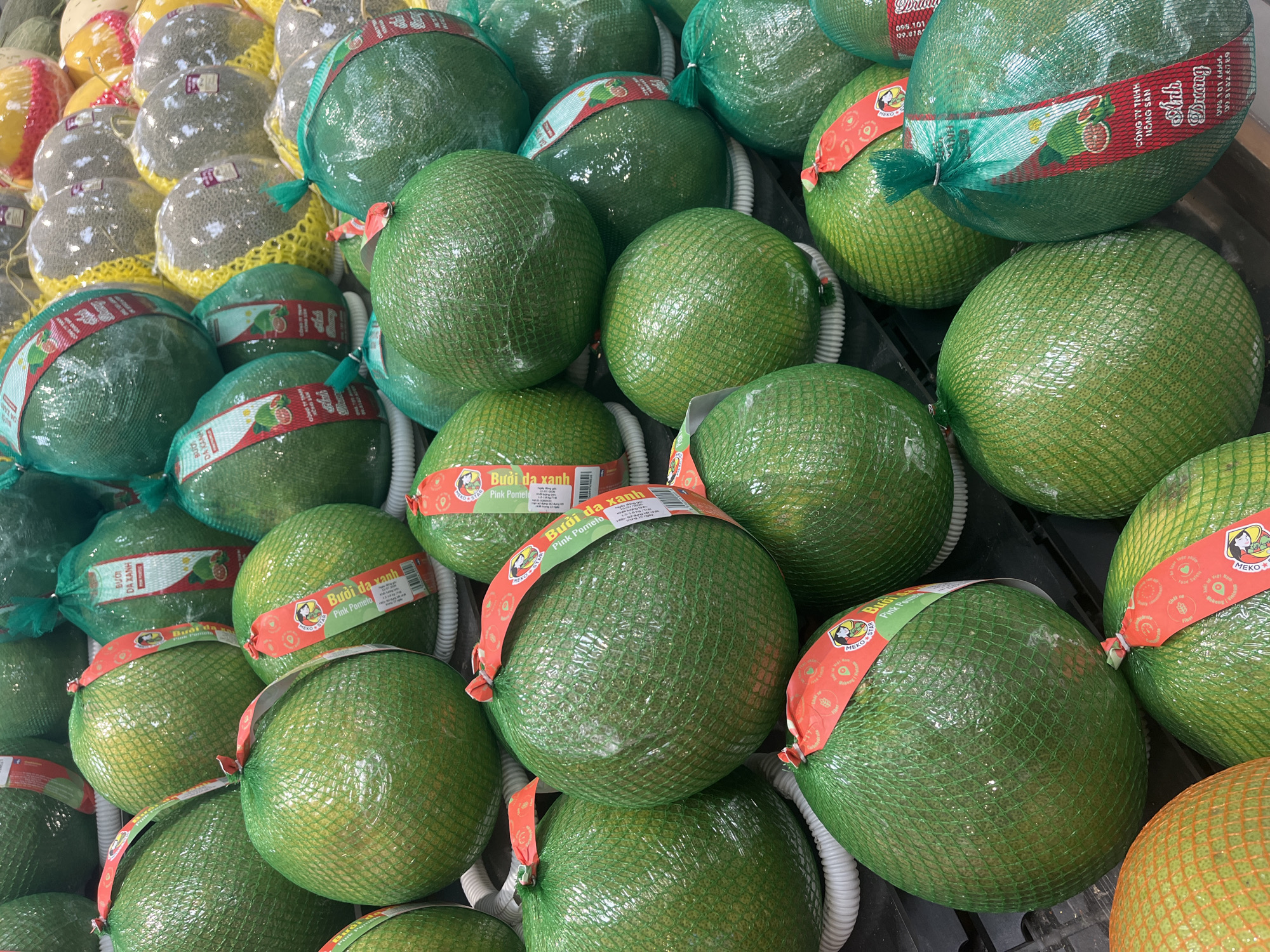 This is a close-up, eye-level shot of a display of pink pomelos for sale in a market. The fruits are large, round, and a vibrant green, each individually wrapped in a green mesh bag. A red and white paper band wraps around each pomelo, featuring Vietnamese text, the brand "MEKO STAR," and illustrations of the fruit and a woman's face. The band also has English text that reads "Pink Pomelo." The fruits are stacked in multiple rows, suggesting a large quantity available. The setting appears to be a market stall or grocery store, possibly in Nha Trang, Vietnam, given the Vietnamese text. The lighting suggests it is daytime, and there are no discernible weather cues. The image focuses solely on the product, with no people or other distracting elements. The emphasis is on the abundance and presentation of the pomelos.