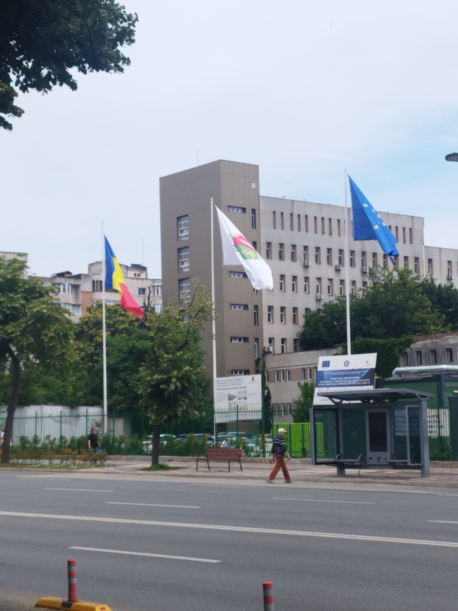 The image captures a street scene in Constanța, Romania, with a modern building, flags, and a bus stop. In the foreground, a multi-lane road with white lane markings is visible, along with a yellow and red traffic delineator.
Behind the road, a bus stop shelter with a metal frame and glass panels stands on the sidewalk. A person in a striped shirt and orange pants is walking away from the bus stop, towards the right side of the image. Another person in a dark shirt is walking away from the bus stop, towards the left side of the image, near a green metal fence.
In the background, a large, beige, multi-story building with numerous windows and air conditioning units dominates the scene. In front of the building, three flags are flying on poles. From left to right, these flags appear to be the Romanian flag (blue, yellow, red), a white flag with a colorful emblem, and the European Union flag (blue with yellow stars). Trees with green foliage are situated between the road and the building, and behind the building, other residential buildings are visible. Two informational signs are placed near the bus stop. The sky is overcast.