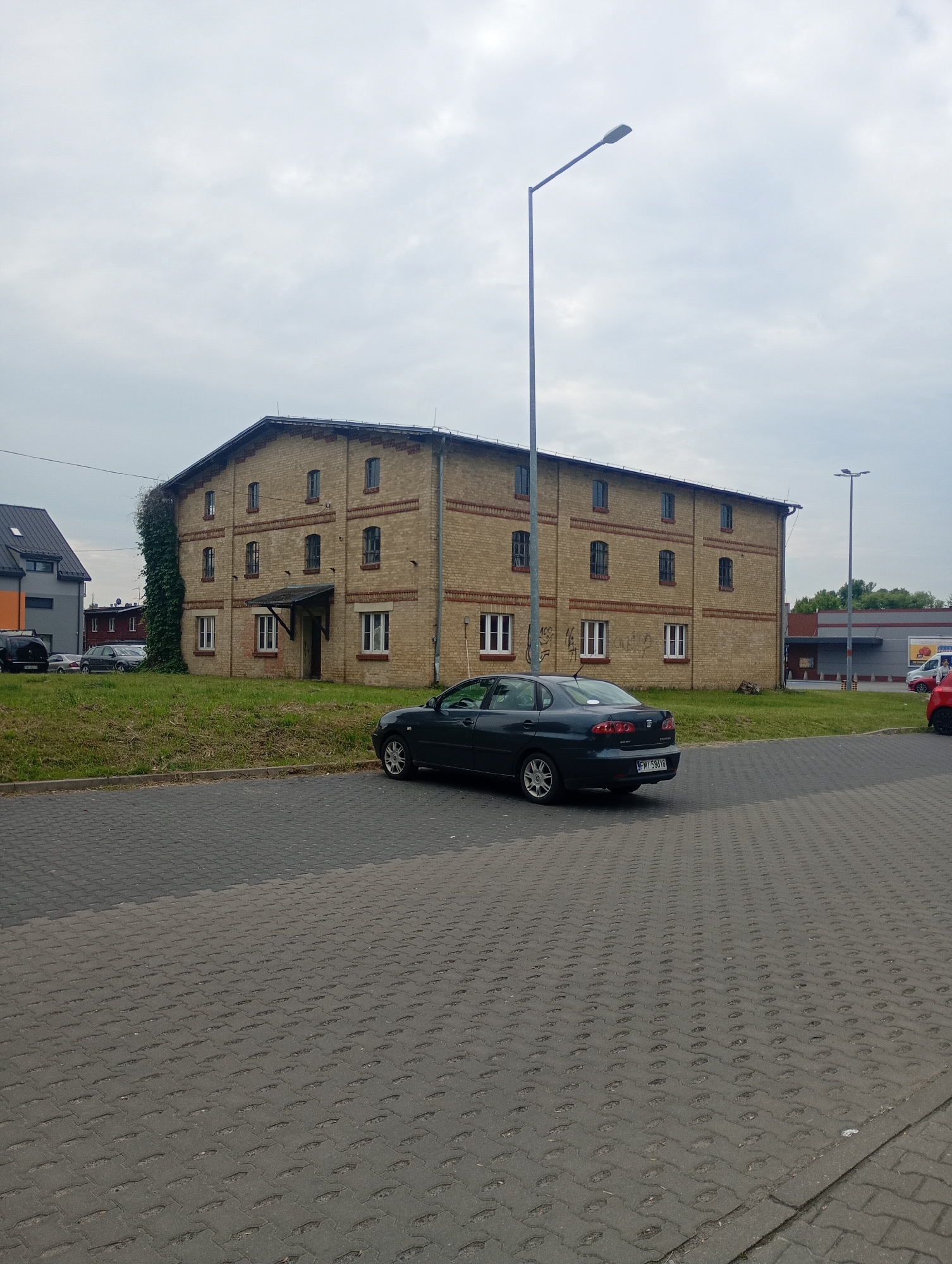 The image depicts a street scene in Międzyrzecz, Poland, on a cloudy day. In the foreground, a dark-colored sedan with the license plate "FM 58618" is parked on a paved lot composed of interlocking gray bricks. To the left of the sedan, a grassy area with some weeds separates the parking lot from a two-story building constructed of yellow brick. This building features numerous windows with white frames on both levels, a dark-colored awning over the entrance, and is partially covered in ivy on the left side.
Further in the background, to the left of the brick building, is a modern building with an orange and gray facade. Beyond the brick building to the right, another building with a gray roof and red accents is visible, possibly a commercial establishment, with a street lamp in front of it. A red vehicle is partially visible to the far right. The sky is overcast, suggesting a temperate climate. No people are visible in the image.