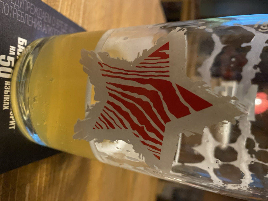 A close-up shot depicts a glass partially filled with a yellow liquid, likely beer. A large, white star design with red, zebra-like stripes is prominently featured on the glass's exterior. The glass also exhibits foam remnants inside, near the rim. Adjacent to the glass, a dark-colored menu or booklet with writing in a Cyrillic-based alphabet is visible. The scene is set on a wooden surface.