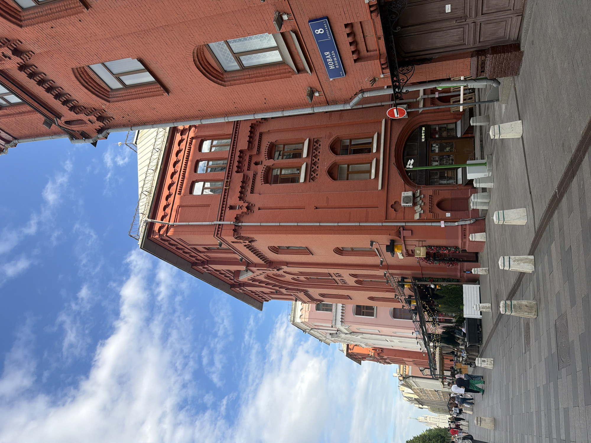 The image depicts a street in Moscow, Russia, with a red brick building on the left side of the frame. A street sign on the building reads "Novaya Square 8". The sky above is blue with white clouds. On the right side, there's a sidewalk paved with gray bricks, and there are concrete bollards along the edge. People are walking on the sidewalk in the distance. There are storefronts and cafes visible along the street.