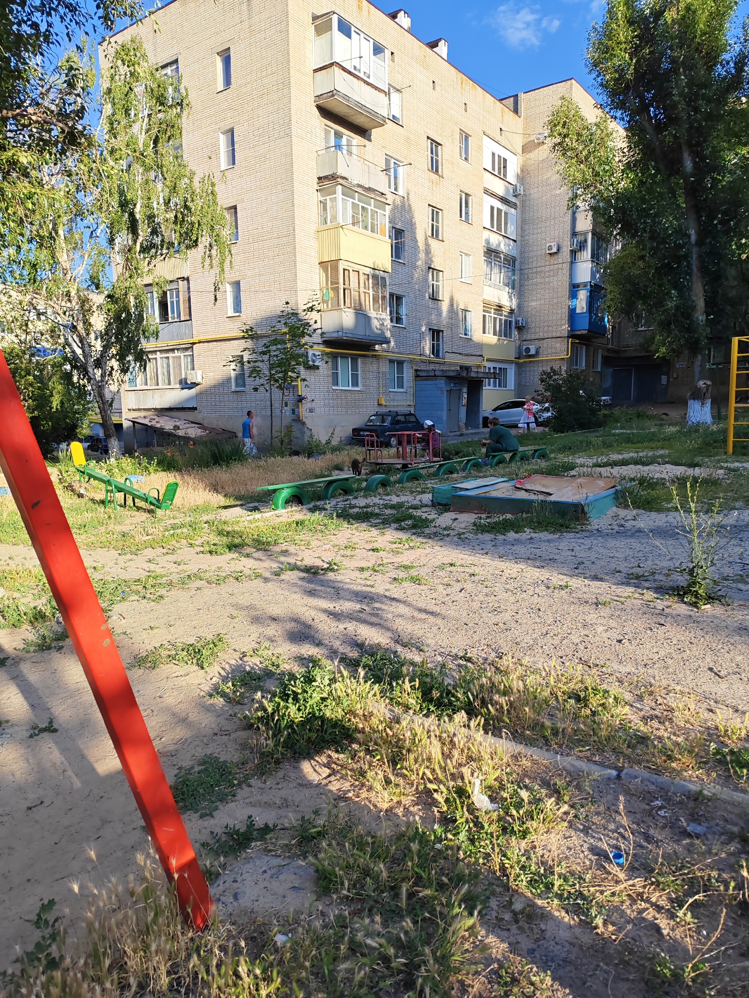 The image shows a residential courtyard in Belaya Kalitva, Russia. A multi-story apartment building constructed of light-colored brick dominates the background. Balconies and windows are visible on its facade. Yellow pipes run horizontally along the building's exterior. Several trees with green foliage are present in the scene, casting shadows on the ground.
In the foreground, there is a playground area with elements made from recycled materials, such as tires. A sandbox with a wooden frame is visible, filled with sand. A seesaw with green seats and a yellow attachment is also present. A large red pole, likely part of a swing set or climbing structure, is prominently featured on the left side of the frame. The ground is covered with sand and patches of dry grass and weeds, suggesting a somewhat unkempt but functional outdoor space.
There are a few people in the scene. One person in a blue shirt and white shorts walks near the building. Another person in a green outfit is seated near the sandbox. A woman in a pink dress is also visible further back. Two black cats are present in the middle ground, near some of the playground equipment.
The lighting suggests it is a sunny day, likely late afternoon or early evening, given the long shadows. The sky is blue with a few wispy clouds. The overall impression is a typical urban residential area, with a focus on the communal outdoor space. There is no visible text in the image.