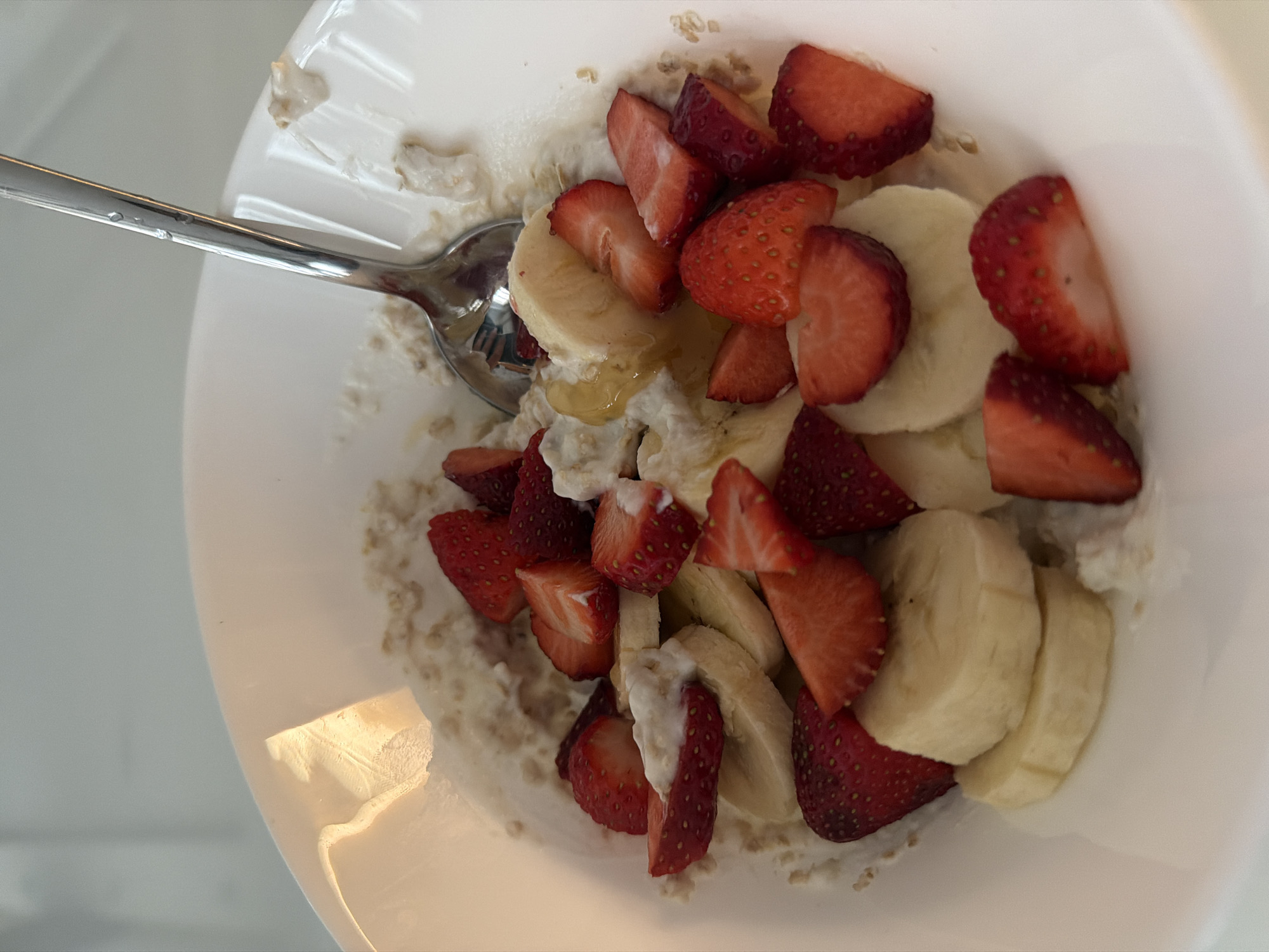 A white bowl contains oatmeal topped with sliced bananas and strawberries. A silver spoon rests inside the bowl. The bowl is situated on a white surface. The location is unspecified, and there is no indication it is in Gabrovo, Bulgaria. There are no people visible in the image.