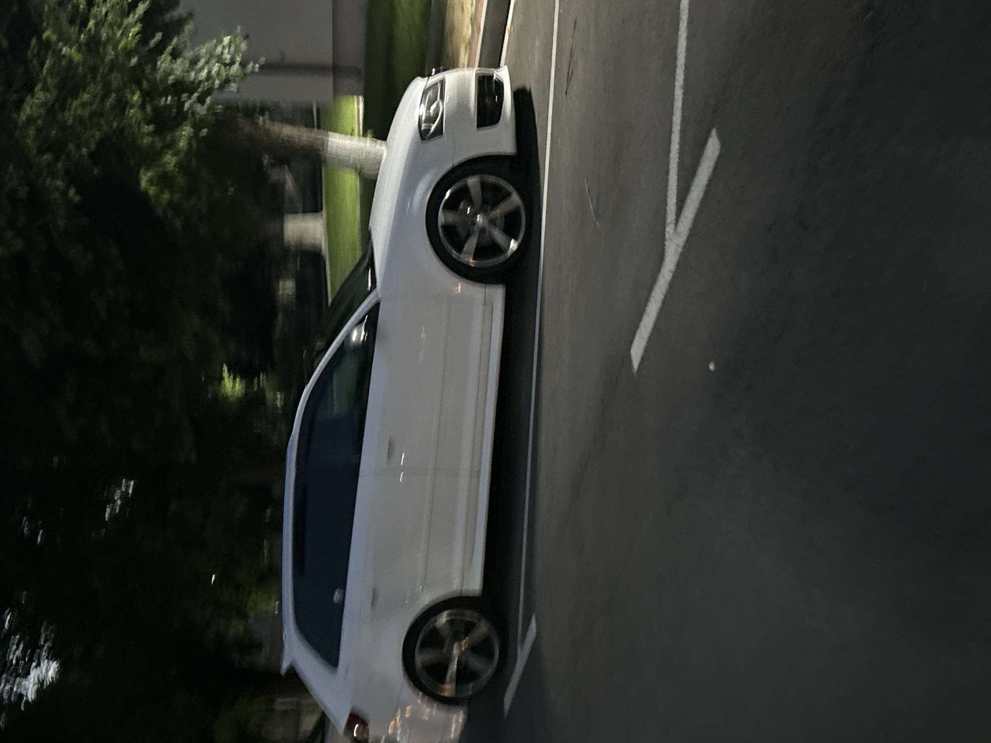 The image displays a white two-door Audi A3 Sportback hatchback parked on an asphalt surface during nighttime in Dzhebel, Bulgaria. The vehicle is oriented with its left side facing the viewer, slightly angled towards the front. It features dark-tinted windows, a subtle roof spoiler, and five-spoke alloy wheels. The front fascia, including the distinctive Audi grille and headlights with integrated LED elements, is visible on the right. The car's ride height appears lowered.
The foreground consists of dark asphalt with white painted lines, indicative of a parking area or road markings. In the background, a strip of illuminated green grass separates the paved area from dense, dark tree foliage. Several tree trunks, appearing light in color, are interspersed within the background. A light-colored building is partially visible on the far right. The scene is illuminated by unseen artificial light sources, creating areas of contrast and shadow. No individuals are present.