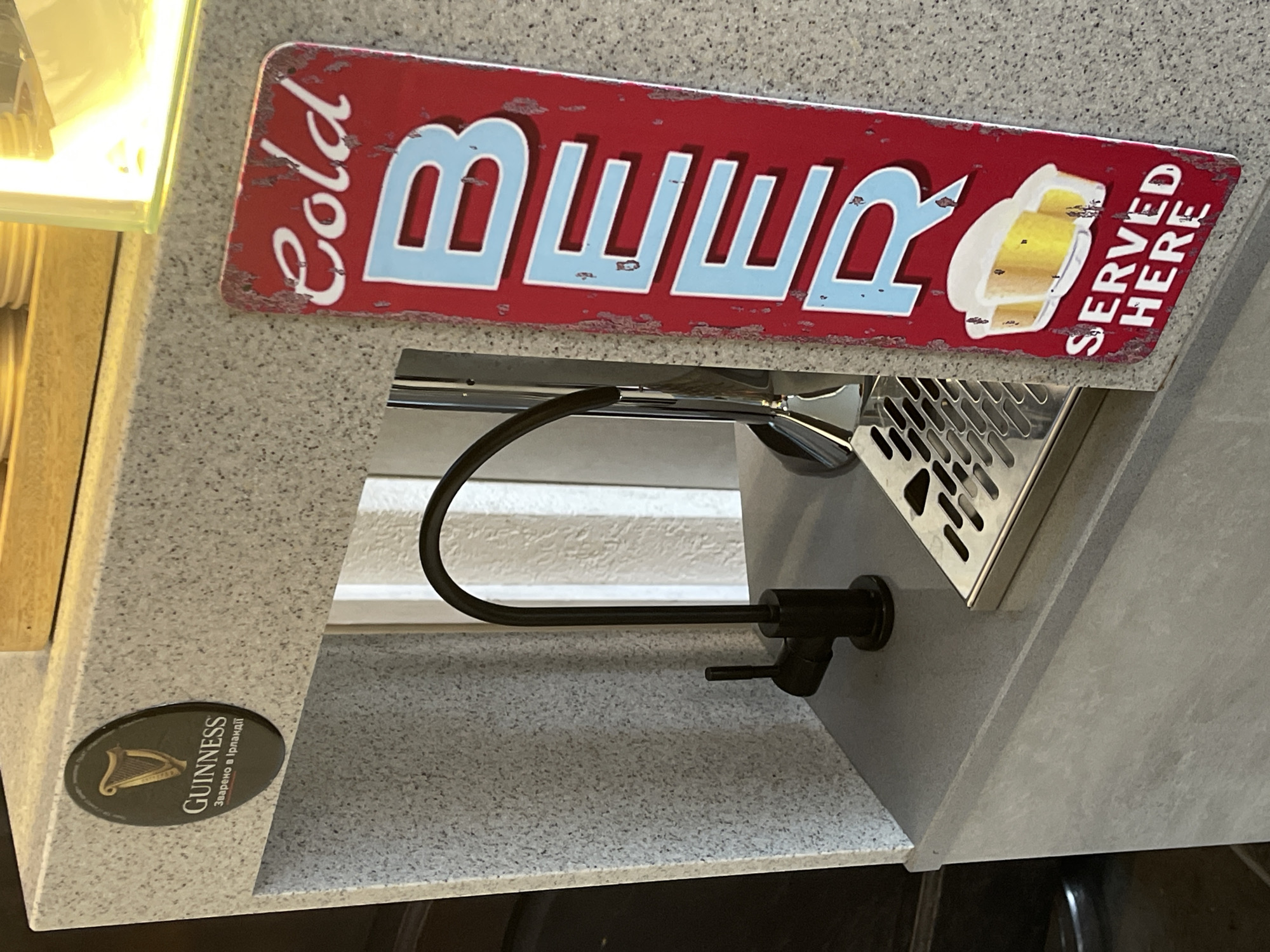A close-up, eye-level shot captures a granite-like surface with a black faucet. To the right of the faucet is a red sign with distressed white and light blue lettering that reads "COLD BEER SERVED HERE" and features an illustration of two glasses of beer. A Guinness logo is visible on the left side of the surface. The background shows a muted, striped pattern. The lighting is bright and appears to be from an overhead source.