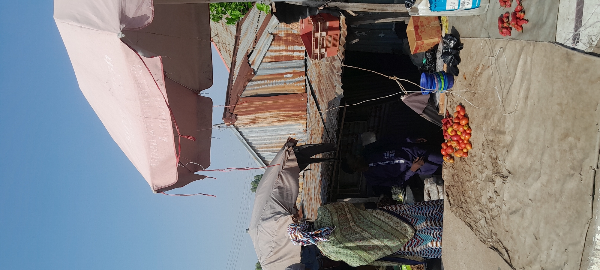This is a medium shot, eye-level, outdoor photograph taken in the city of Jalingo, Nigeria. The scene depicts a bustling market with two individuals. In the foreground, a large pile of red tomatoes is displayed on a tarp. To the left, a person wearing a colorful patterned headwrap and a green, patterned robe stands facing away from the camera, towards a stall. To the right of the tomatoes, a man in a dark blue, patterned robe is seated, looking towards the tomatoes. Behind them, a market stall is constructed with corrugated metal sheeting, showing signs of rust and age. A large pink umbrella shades part of the scene from the sun. In the background, more market stalls and structures are visible, along with a clear blue sky. The overall impression is of a typical day at a Nigerian open-air market.