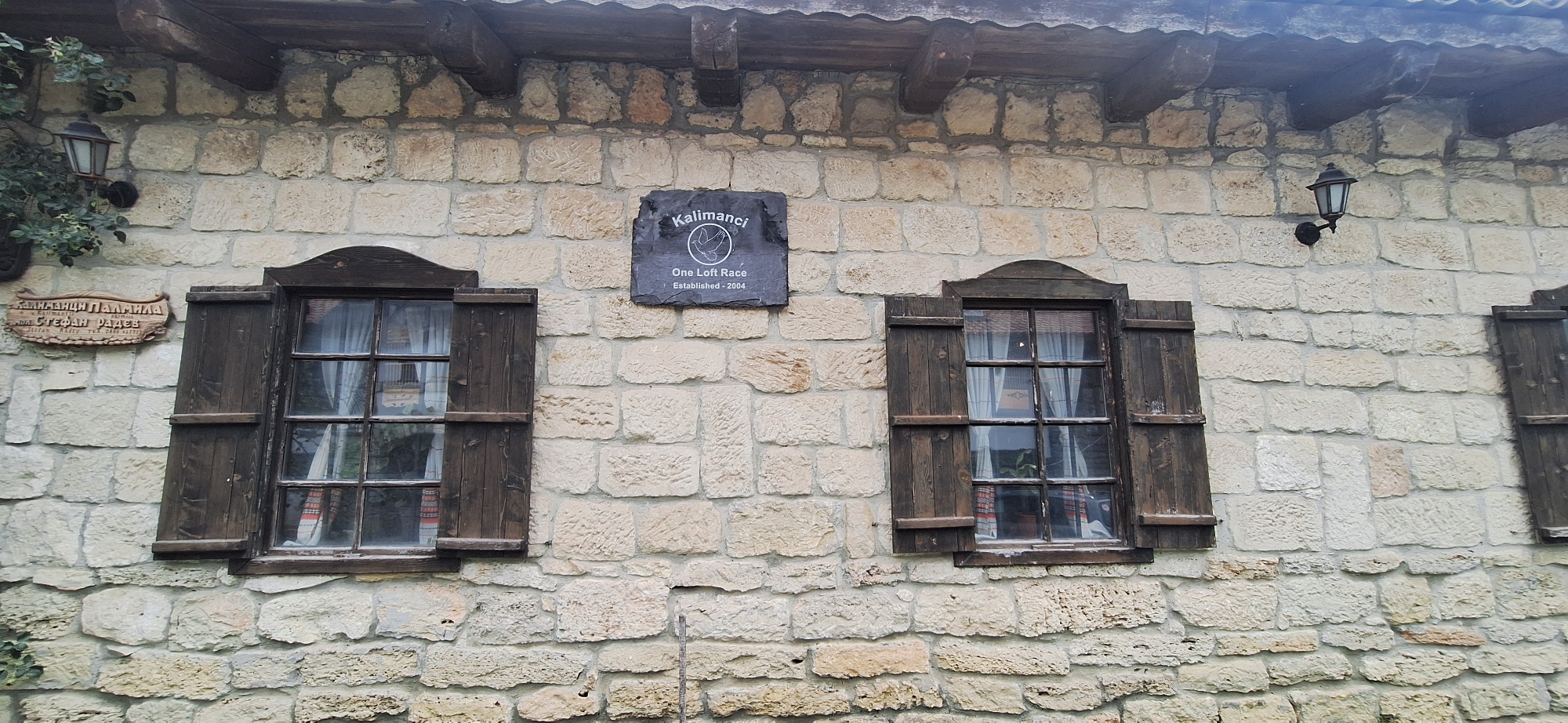 The image shows a stone wall of a building with two windows. One window is on the left and the other is on the right. Both windows have wooden shutters and are partially open. Above the windows, there is a dark sign with white text that reads "Kalimanci One Loft Race Established - 2004". To the left of the sign, there is another sign with text in Bulgarian. The wall is made of roughly cut stones. There are two outdoor lamps attached to the wall, one to the left and one to the right. Green foliage is visible to the left of the left window. The overall scene appears to be an exterior shot of a building in Kalimantsi, Bulgaria.