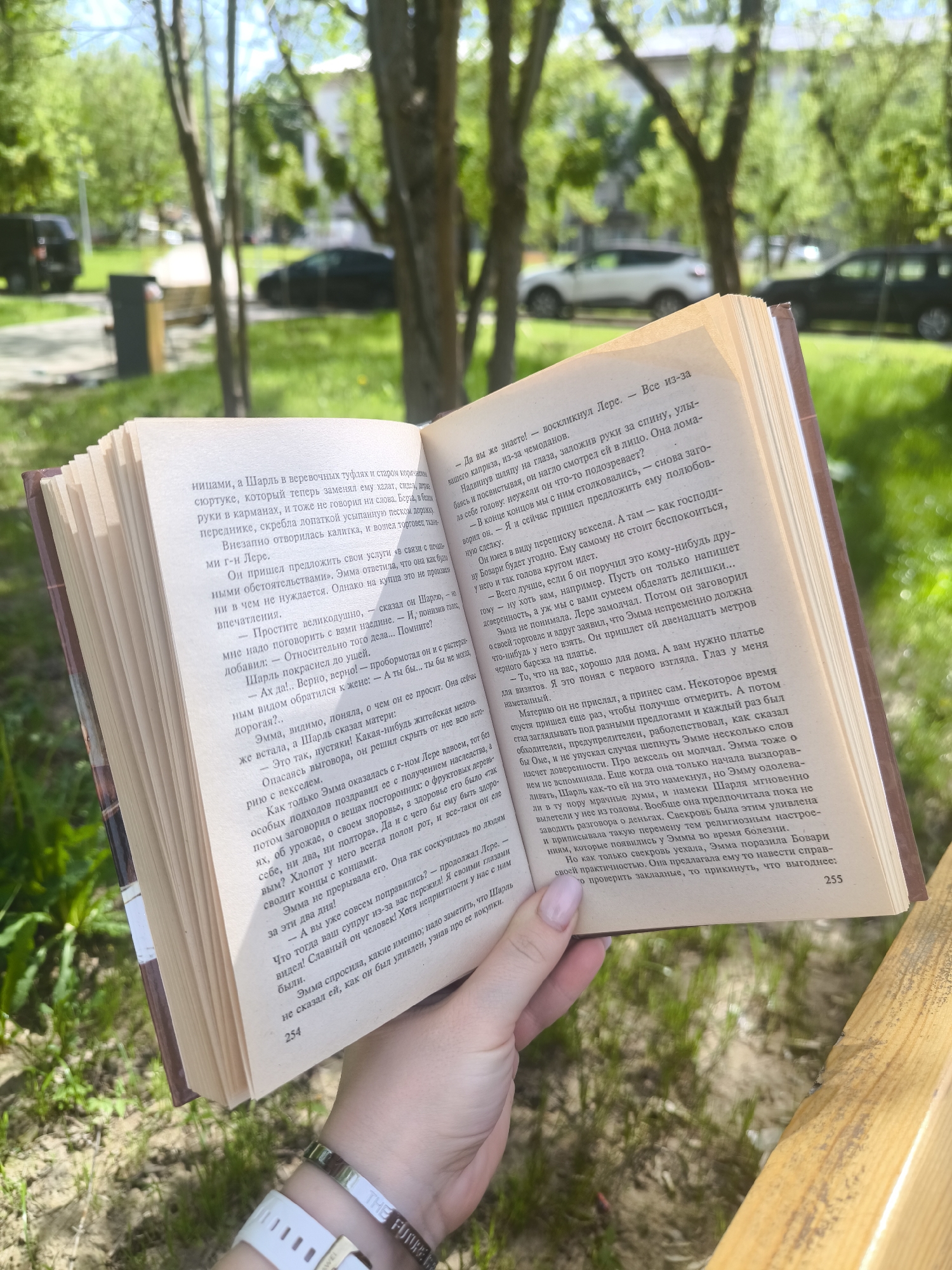 The image shows a person holding a book open, with the text in Russian visible on the pages. The photo is taken outdoors with a blurred background showing a park-like setting with trees and what appears to be a street with parked cars. The focus is on the book, which is resting in the holder's hands. The person's left wrist, adorned with a watch and bracelet, is also visible. The book is positioned to face the camera and the natural light casts soft shadows on the pages. Based on the text on the pages, the location in the city of Fryazino, Russia could be linked to any person holding an open book.