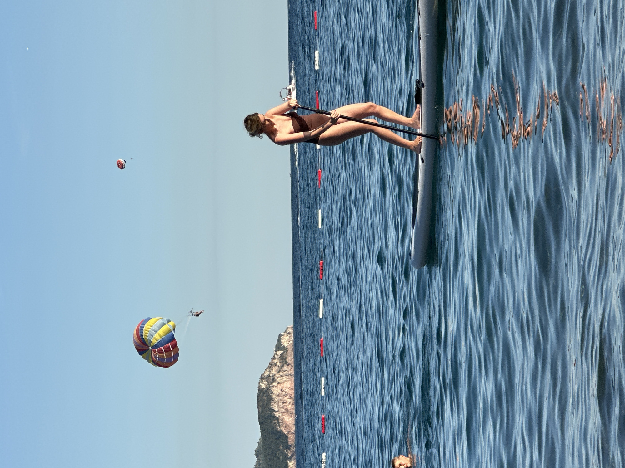 This is a horizontal, eye-level, outdoor photograph of a beach scene in Boreti, Montenegro. In the foreground, a woman is paddleboarding on the water, holding a paddle. Her body is angled slightly away from the camera, and she appears to be looking down at the water. She is wearing a brown swimsuit. The paddleboard is gray and white. In the mid-ground, another person is swimming in the water, their head and shoulders visible. There are buoys in the water separating swimming areas. In the background, a large, rocky island with trees is visible. Above the water, a colorful parasail is in the sky, with a person attached to it. Further in the distance, another parasail is visible, smaller and further away. The sky is clear and blue. The water is calm with small waves.