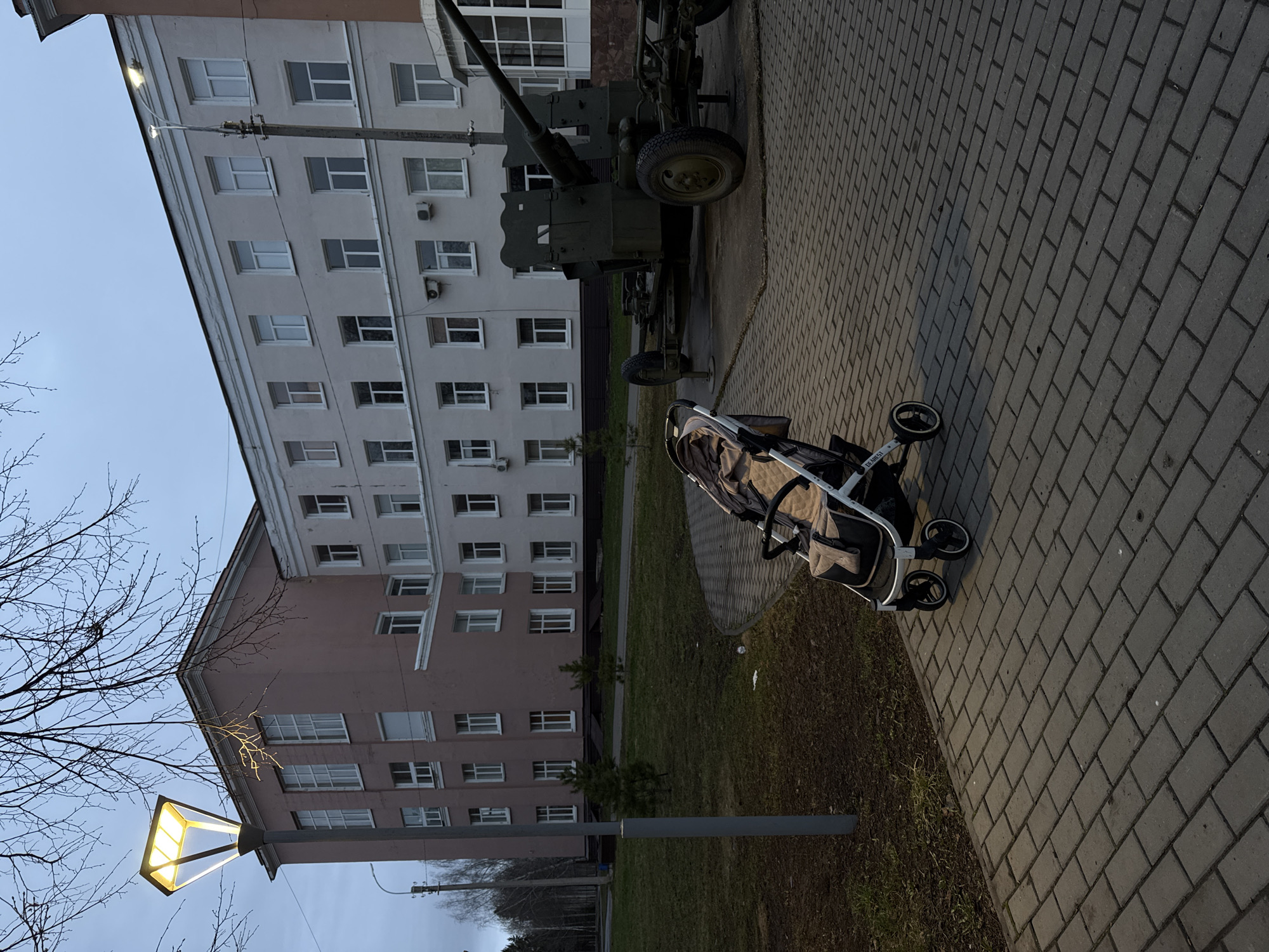 The image shows a street scene in Izhevsk, Russia. An anti-aircraft gun stands on the sidewalk beside a brick-paved area near an apartment building. A baby stroller sits nearby on the brick path. A streetlight illuminates the foreground. The sky is overcast. There are no visible people.