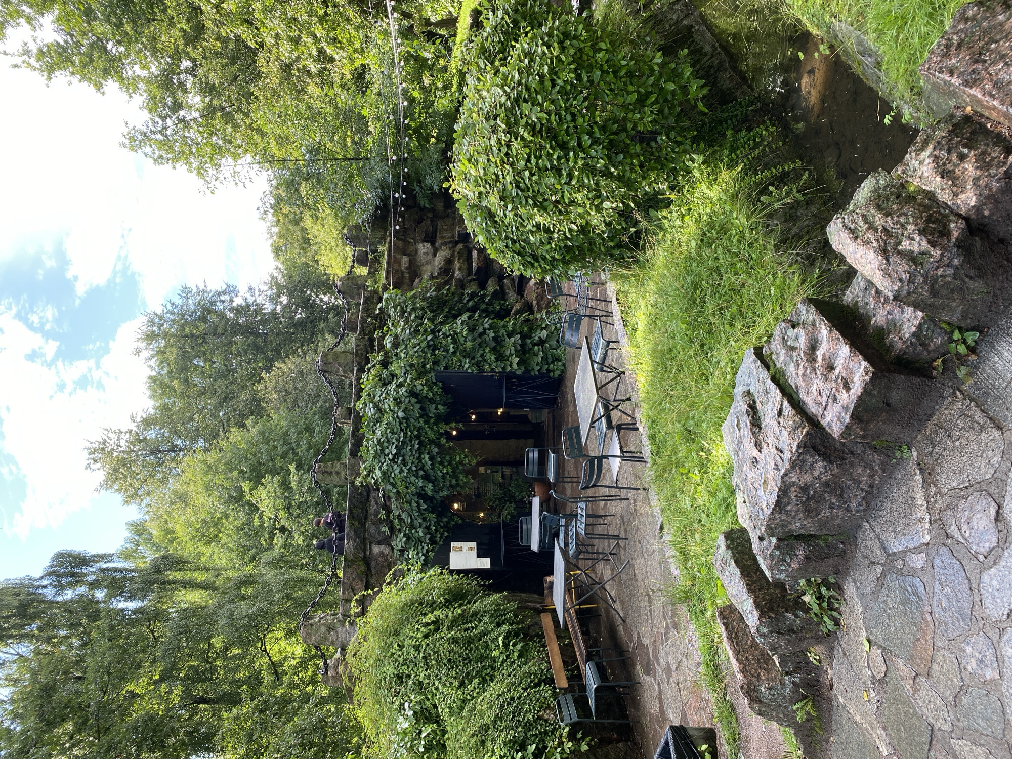The image shows an outdoor seating area of a cafe situated in a park-like setting in Saint Petersburg, Russia. In the foreground, large granite boulders line a stone pathway leading to the cafe's entrance. Beyond the pathway, several metal tables and chairs are arranged on a paved patio. A wooden bench is visible to the left of the patio. The cafe's entrance is integrated into a stone structure overgrown with ivy. String lights are hung above the seating area, and lush green foliage surrounds the entire scene, including a small stream on the right. Two individuals are visible in the background, standing on top of the stone structure. The sky above is partly cloudy.