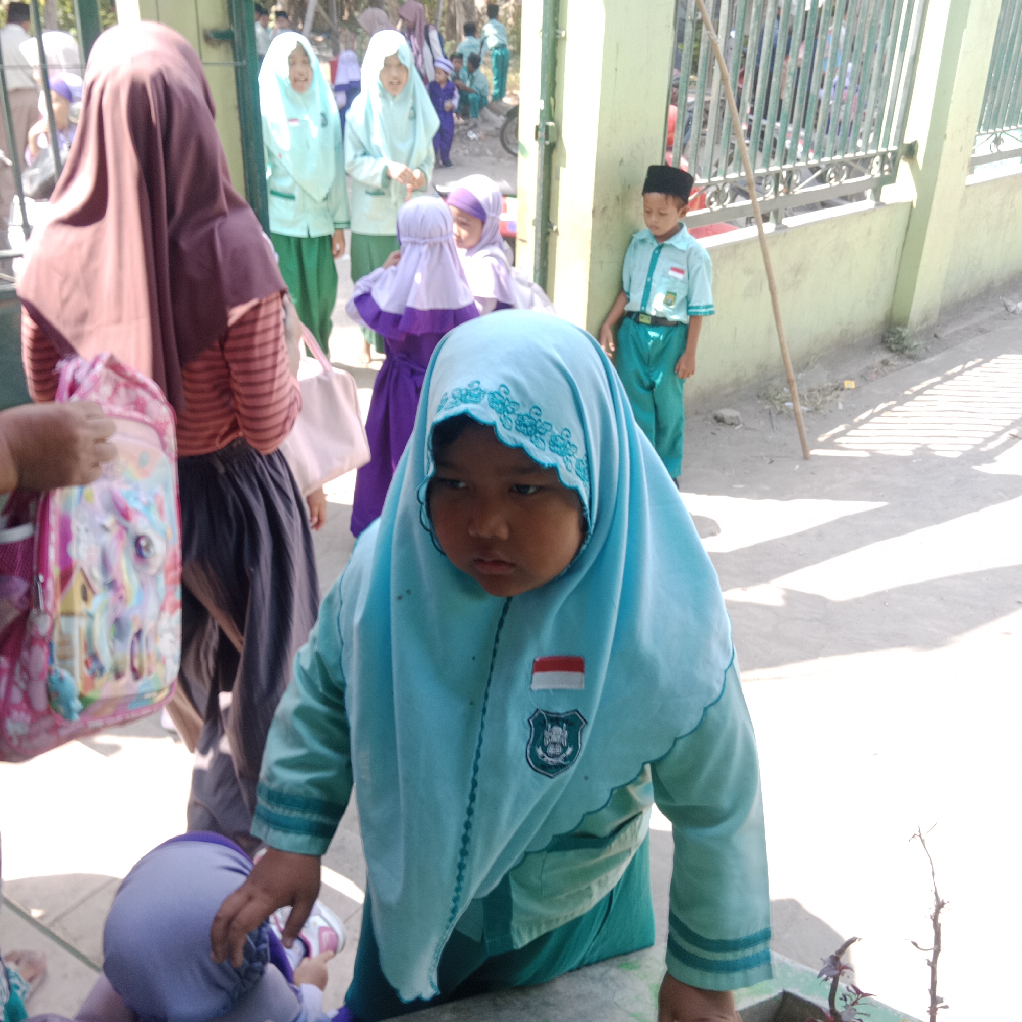 The image depicts a group of children, likely students, standing outside what appears to be a school building. The scene is set in the city of Tegowanu Kulon, Indonesia, as indicated by the presence of individuals wearing school uniforms with Indonesian flags on their apparel. In the foreground, a young child wearing a light blue hijab with decorative trim is looking down with a pensive expression. This child is positioned near a wall and is partially obscuring another child wearing a purple hijab, who is seated and being helped by an adult's hand. Behind them, several other children are visible, some in similar school uniforms with light blue or green tops and green pants. One child, a boy, stands to the right wearing a light blue shirt with a crest and a black cap, while another child in a mint green hijab is in the background. A woman in a maroon hijab is seen in the left foreground, holding a pink backpack adorned with colorful cartoon characters. The environment suggests an outdoor setting with a light-colored wall and a metal gate in the background, along with a sandy or dirt ground. The lighting indicates a sunny day with distinct shadows cast on the ground.