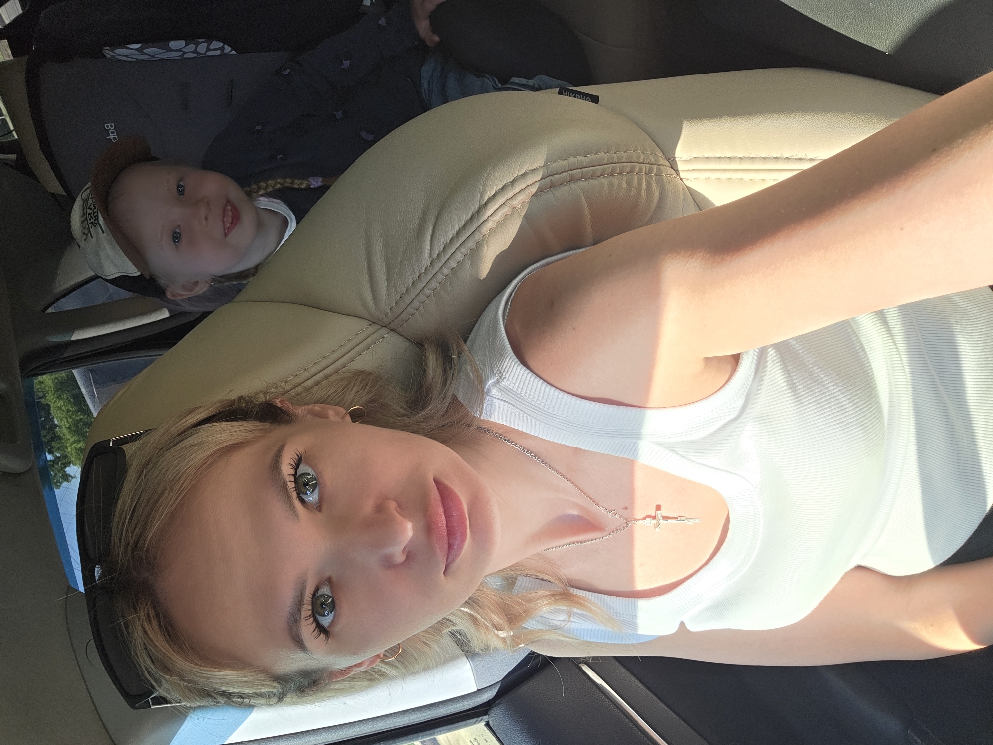 A selfie of a woman with light skin and light blond hair is taken inside a car. She wears a white top and a necklace with a cross pendant. Her eyes are light, and she has pink lipstick on. Sunglasses are partially visible on her left side. A child with light hair and a baseball cap lies in a car seat in the background. The car's interior is beige. The location is unknown, and no information suggests this image was taken in Chaikovsky, Russia.