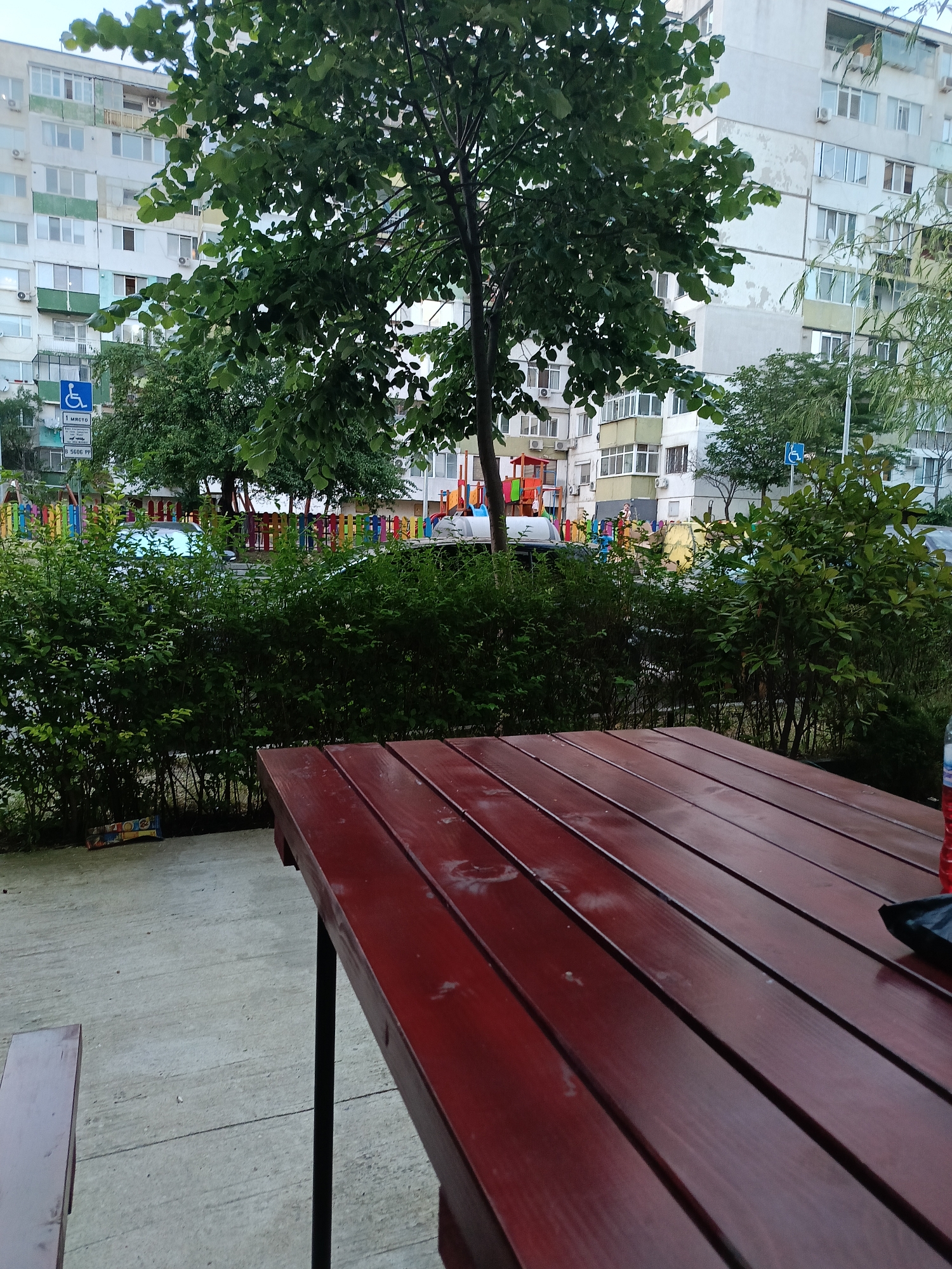 The image shows an outdoor seating area in a residential neighborhood in Varna, Bulgaria. In the foreground, a red wooden picnic table is partially visible, with a concrete ground beneath it. Behind the table, there is a dense green hedge. Further in the background, a children's playground with colorful fencing and equipment can be seen. Residential buildings, characteristic of Varna's urban landscape, rise behind the playground. A sign indicating a disabled parking spot is also visible on the left side of the image. The overall scene depicts a common urban amenity in a Bulgarian city, suggesting a public or semi-public space for recreation and relaxation.