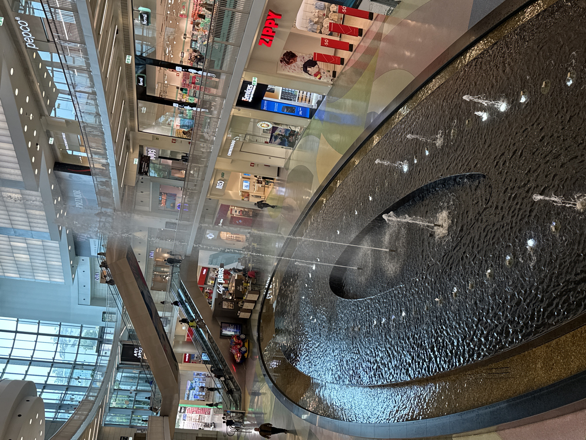 The image captures an interior view of NorteShopping in Matosinhos, Porto, Portugal. Dominating the scene is a large, oval-shaped water fountain with multiple jets spraying water upwards. The fountain’s edge is lined with dark stone.
In the background, various retail stores span multiple levels, including ZARA, PRIMOR, pepco, and ZIPPY. Escalators are visible, connecting different levels of the shopping center, with people using them. Several people walk through the shopping center. The building features a high ceiling and large windows.