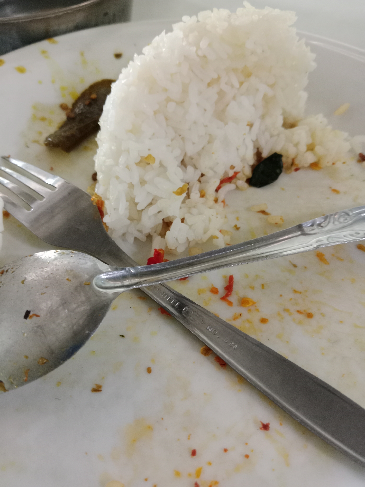 The image shows a white plate with leftover rice and bits of food scattered across its surface. A fork and a spoon are resting on the plate, partially obscuring the food remnants. The fork is positioned above the spoon, with their handles extending towards the bottom of the frame. The plate appears to be in a dining setting, likely a restaurant or a home. The lighting suggests an indoor environment. There are no people visible in the image. The overall scene depicts the aftermath of a meal. The location in West Jakarta, Indonesia, is inferred from the context provided.