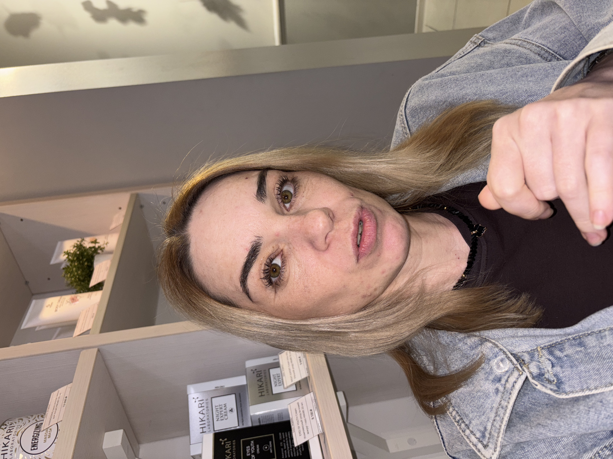 A medium shot captures a fair-skinned woman with blonde hair and green eyes, looking directly at the camera. She is wearing a denim jacket over a dark brown shirt, and a black beaded necklace. Her expression is neutral, with her mouth slightly open. The background consists of shelves displaying various cosmetic products, suggesting the setting is a store. To the left, a shelf holds products from the "HIKARI LABORATORIES" brand, including an "Energizing Massa Mask" and "Eyes of Youth Cream." Another shelf displays "Night Expert Cream." The lighting is soft, creating a slightly diffused atmosphere. The image appears to be taken indoors, possibly in a retail environment within Ladozhskoe Ozero, Russia.