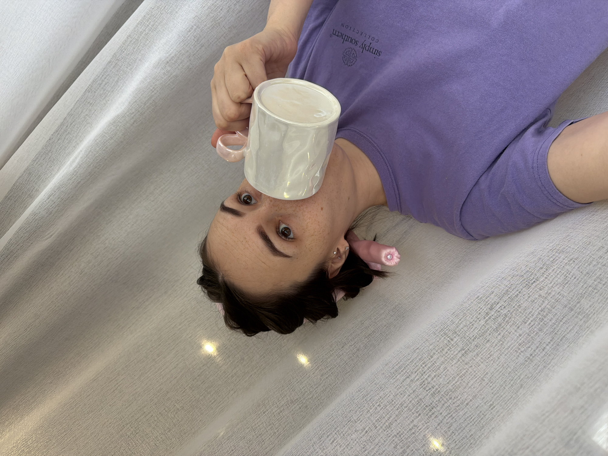 The image depicts a person, upside down, holding a white iridescent mug with a pink handle up to their face. The person is wearing a purple shirt with a "Simply Southern Collection" logo on the upper left. Their hair is dark, with pink accessories. The background is a wrinkled, light-colored fabric, possibly a curtain or sheet. The setting appears to be indoors. There is no definitive information to confirm the location as Chaikovsky, Russia.