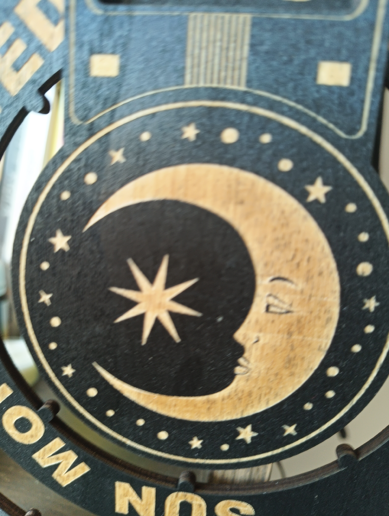 This is a close-up, vertically oriented, indoor photograph of a decorative wooden ornament featuring celestial imagery. The ornament has a dark blue background with a circular motif. Within this circle is a crescent moon, depicted in wood grain, with a stylized face. The moon is surrounded by small wooden stars and dots. The ornament appears to be part of a larger piece, with some text visible at the bottom that reads "SUN MO... NOS". The overall impression is of a handcrafted, possibly mystical or astrological-themed item. No specific location within Karnobat, Bulgaria can be determined from the image.