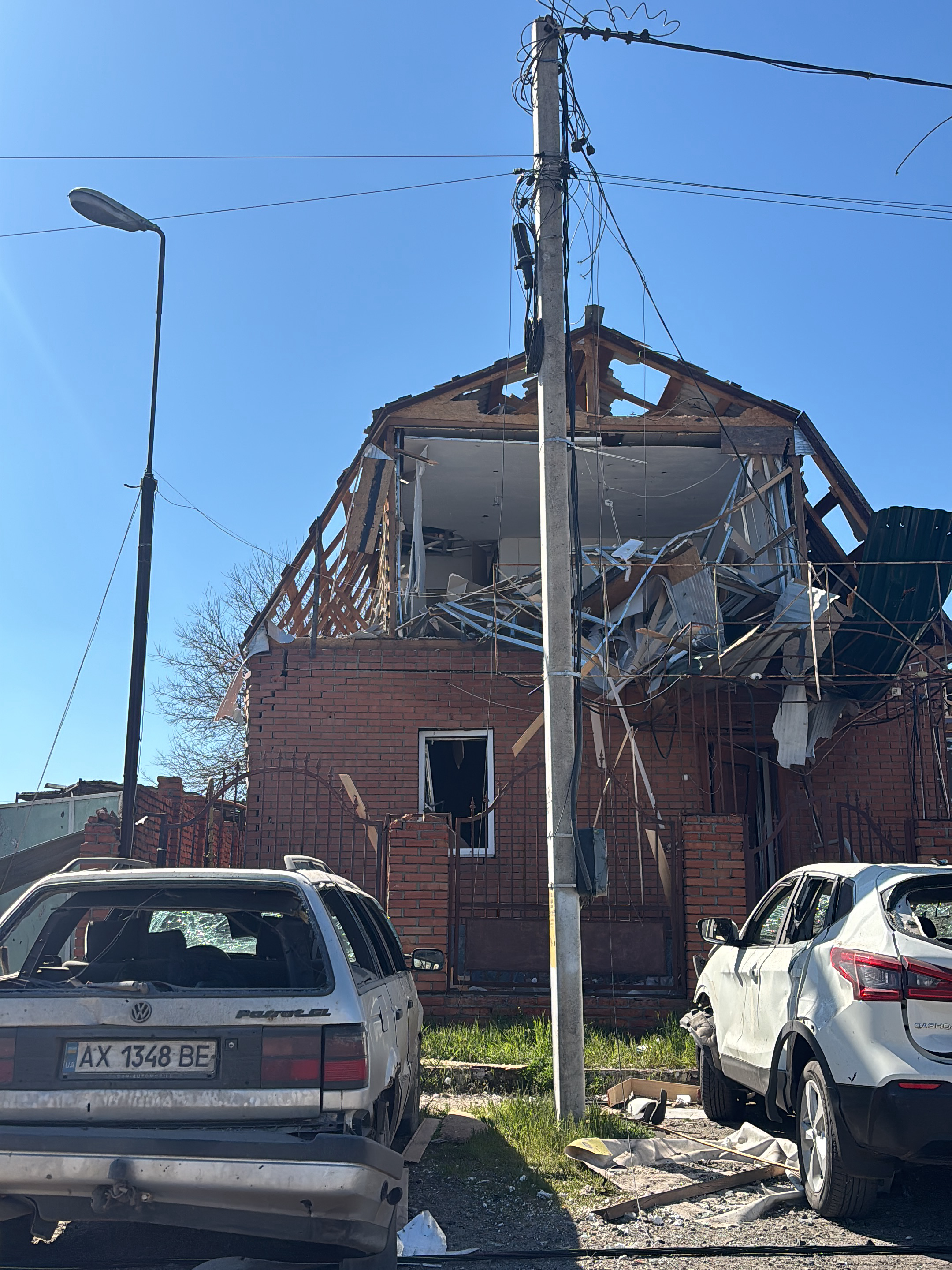 The image shows a severely damaged residential area in Vysokyi, Ukraine. A two-story brick house is heavily damaged, with a collapsed roof exposing the interior. Debris is scattered around the property. Two cars parked in front of the house are also damaged, with broken windows and visible damage to their bodies. A utility pole stands near the house, with power lines extending across the top. A street light is visible on the left side of the image. The sky is clear and blue. The car license plate visible shows registration in Ukraine.