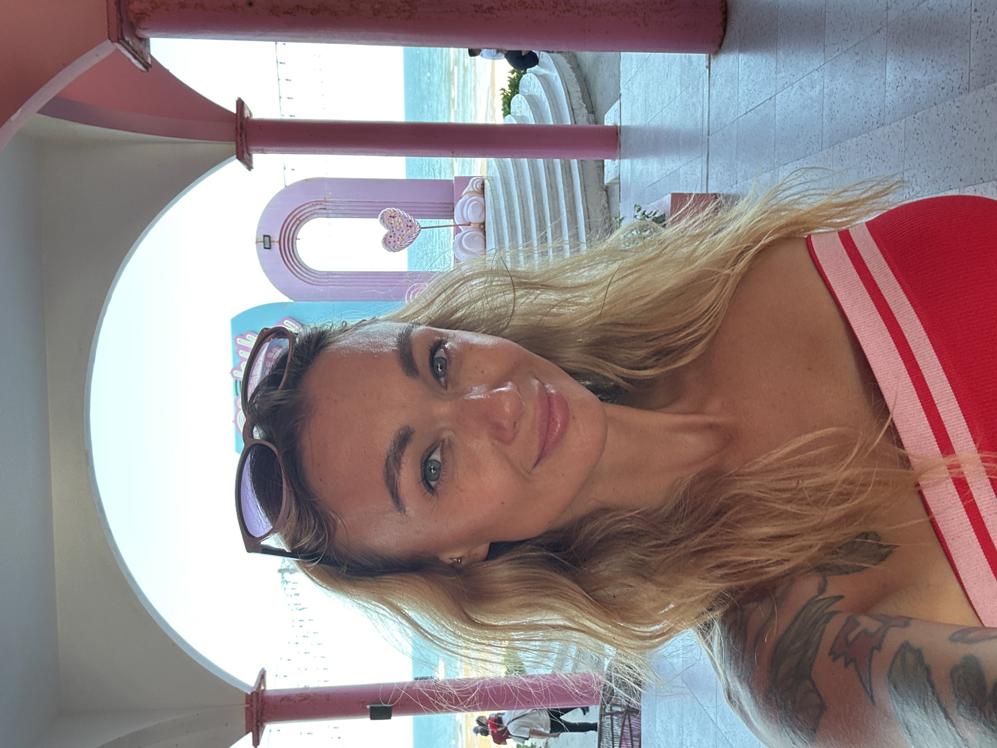 A woman with blonde hair pulled back and sunglasses perched on her head takes a selfie in front of a vibrant pink structure with arched openings. She is wearing a red and white striped strapless top. In the background, the blue ocean is visible, and there are white steps leading to an archway decorated with a pink heart lollipop. To the left, a few people are walking near the water. The overall setting appears to be a beachfront resort or attraction in Tien Thanh Ward, Vietnam.