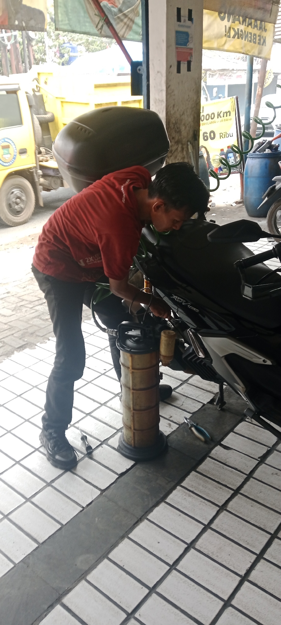 In Suka Manah, Indonesia, a mechanic in a red shirt and black pants works on a black motorcycle parked on a tiled floor. He is using a yellow oil drain pan with a transparent cylinder to collect fluid from the motorcycle's engine. The mechanic is positioned to the left of the motorcycle, bending over to access the engine area. Behind the motorcycle, a black storage box is attached to its rear. To the left of the scene, a yellow truck is partially visible, with its wheel and a portion of its body showing. In the background, there are various objects including blue barrels, a sign with text, and some vegetation. The overall setting appears to be an outdoor service area or workshop.