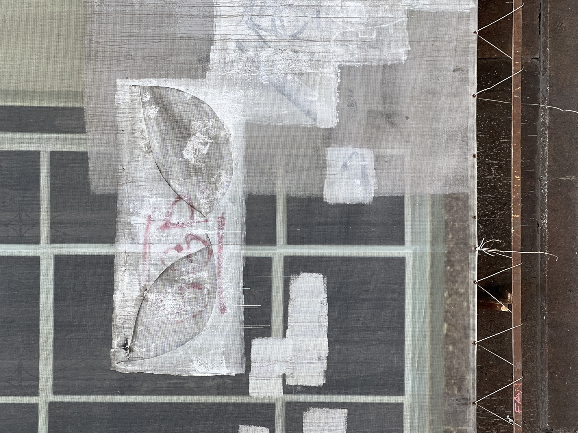 The image depicts a close-up view of an urban surface, possibly a window covered with a translucent mesh screen, creating a textured and somewhat abstract composition. The scene is dominated by a grid pattern, likely from window frames, visible behind a light gray, fine-mesh fabric that covers most of the frame. This mesh is adorned with various markings: broad strokes of white paint, some appearing as fresh rectangular blocks, others as smudged or weathered patches.
Prominently featured on the left side of the mesh are stylized, abstract shapes in a reddish-pink hue, resembling graffiti or faded markings. One distinct element within these markings is an irregular, leaf-like or organic shape. The mesh material itself shows wrinkles and varying degrees of transparency, allowing the dark grid pattern behind it to peek through.
On the far right, a vertical section reveals a dark, rusted metal beam or frame, to which the mesh screen is attached. White wires are laced in a zigzag pattern through small eyelets along the mesh's edge, securing it to the metal structure. To the right of the metal beam, a darker, roughly textured wall, possibly concrete, completes the background.
The overall impression is one of urban decay, abstract art, or a temporary construction covering. The lighting suggests daytime, with no harsh shadows.
Visible text includes the letters "FAN" clearly stenciled or painted in reddish-pink on the lower part of the rusty metal beam on the right. Some of the abstract red markings on the mesh might contain indistinct letters, but they are too obscure to decipher confidently.