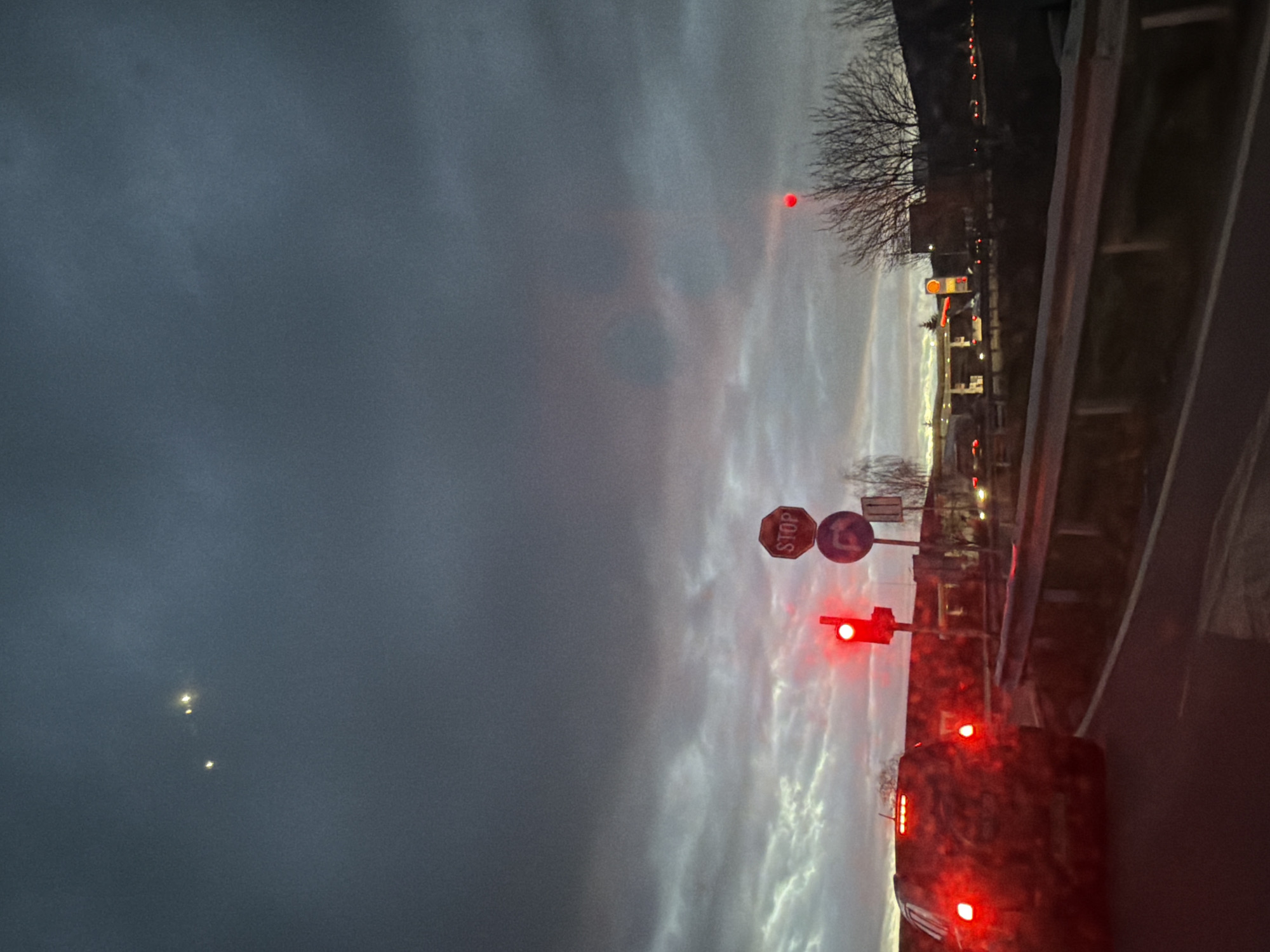 The image shows a street scene taken from a moving vehicle. It is dusk or dawn, and the sky is overcast. In the foreground, the rear of a vehicle is visible, with its red taillights illuminated. A traffic light is also illuminated in red, indicating a stop. A stop sign is present, along with a right-turn sign. Beyond the immediate road area, buildings and trees are visible against the skyline. The location is Dragichevo, Bulgaria.