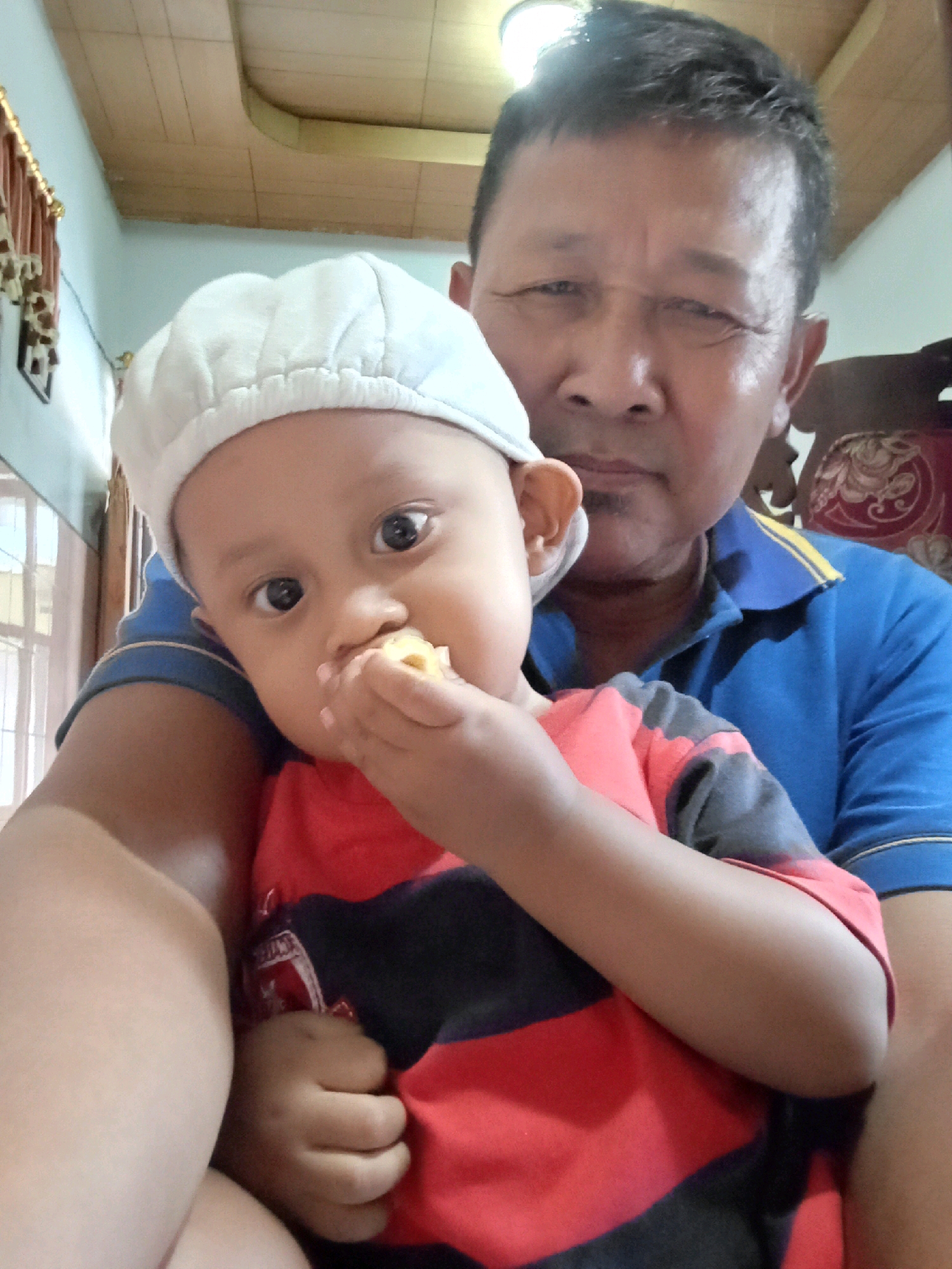 A close-up, indoor, selfie-style shot captures a man and a child in what appears to be a residential setting. The man, in the foreground and slightly out of focus, wears a blue polo shirt with yellow trim. He is holding a young child, who is the primary subject and is in sharp focus. The child wears a white cap and a red and black striped shirt. The child's face is turned towards the camera, with their mouth open and a piece of food being held to it. The child's eyes are wide and directed at the camera. The background suggests an interior space with a slatted wooden ceiling, light blue walls, and a window with blinds visible on the left. A patterned upholstery of a chair is visible behind the man on the right. The lighting is bright, likely from an overhead source. The image is consistent with a personal photograph taken within a home in Kebonagung, Indonesia.