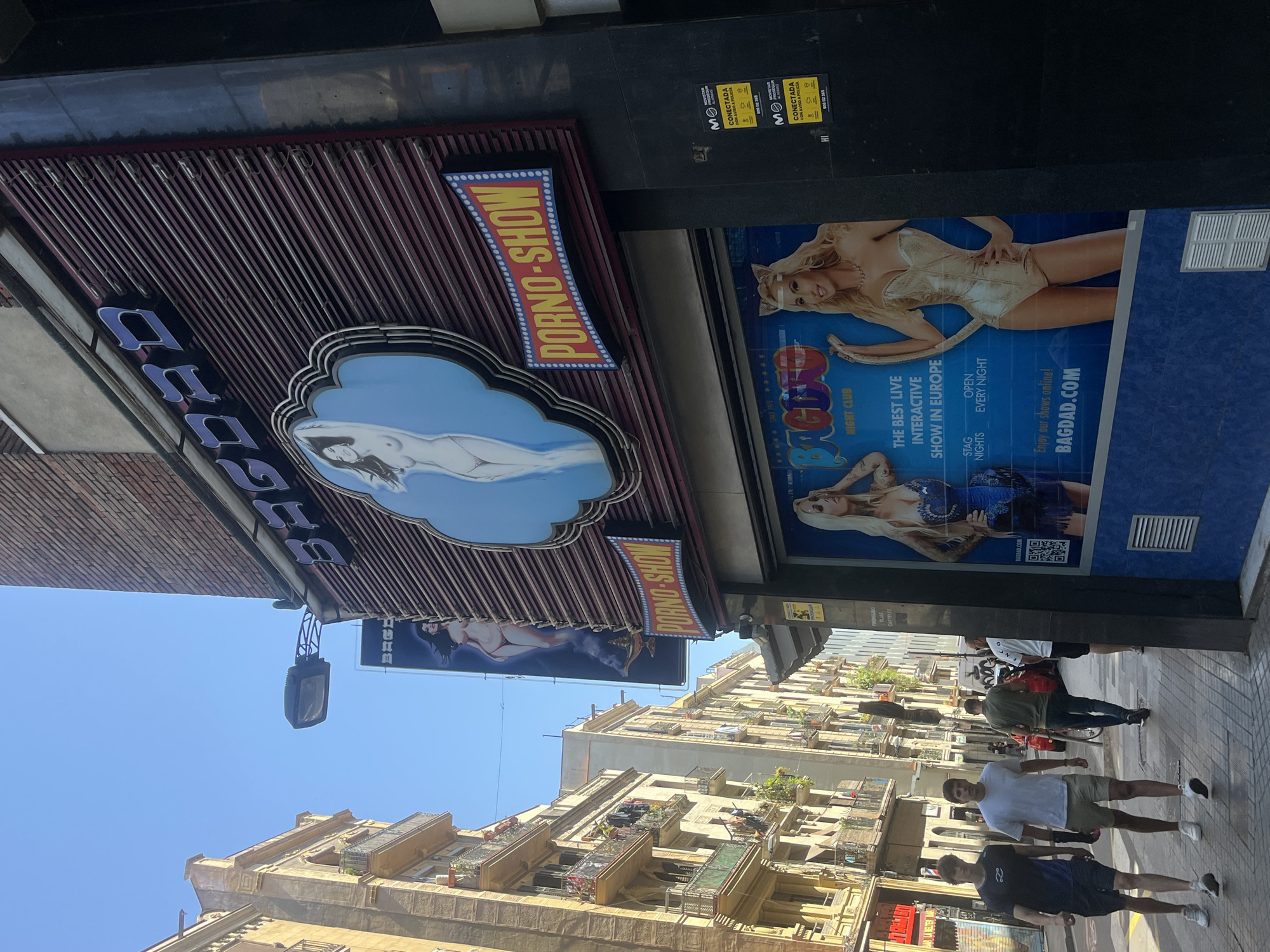 The image captures a street scene in Barcelona, Spain, focusing on the facade of a building with prominent signage for "BAGDAD" and "PORNO-SHOW." The building appears to be a commercial establishment, likely a nightclub or adult entertainment venue, given the explicit nature of the signs and the imagery displayed on the posters.
On the upper part of the facade, the word "BAGDAD" is illuminated in large, stylized letters, with a blue neon outline. Below this, a circular sign features a graphic of a nude female figure. To the right of this, a rectangular sign in red and yellow reads "PORNO-SHOW." Further down, a large poster showcases two women in suggestive attire, with text indicating "BAGDAD NIGHT CLUB" and "THE BEST LIVE INTERACTIVE SHOW IN EUROPE." The poster also includes the website "BAGDAD.COM."
The street itself is paved, with a sidewalk running alongside the building. Several people are visible walking on the sidewalk. On the left side of the image, two young men are walking towards the viewer, dressed casually. Further down the street, other individuals are seen walking away from the camera. The architecture of the surrounding buildings suggests a dense urban environment, with apartment buildings featuring balconies and laundry hanging out to dry. The sky is clear and blue, indicating a sunny day. The overall impression is that of a bustling city street with a clear emphasis on the commercial entertainment offerings of the "BAGDAD" establishment.