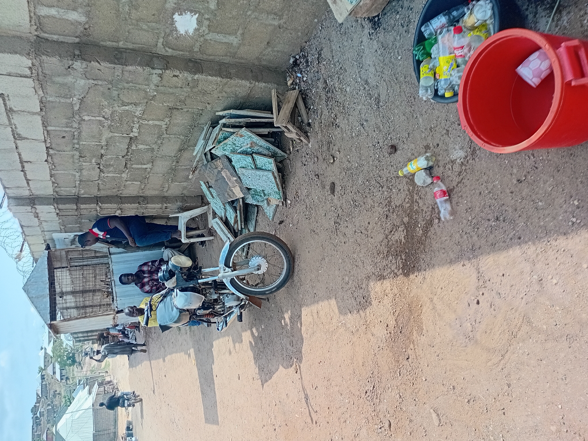 The image captures an outdoor scene in Lokoja, Nigeria, characterized by an unpaved, dusty environment alongside a concrete block wall. On the left side, the rough-textured wall forms a boundary, with stacks of what appear to be broken green-patterned tiles and wooden planks piled against its base.
Several people are present. A man in a dark blue shirt with red collar and sleeve details stands near the wall. Closer to the foreground, a silver motorcycle with chrome accents and a black seat is parked. A man in a yellow shirt is seated on the motorcycle, while another man in a plaid shirt sits on a white plastic chair directly behind it. In the background, beyond these individuals, other people and structures, including buildings with corrugated metal roofs, are visible. Barbed wire is also seen on a structure to the left.
On the dusty ground to the right, there's evidence of discarded items. A black bucket is filled with numerous clear plastic beverage bottles, many containing liquid or residue, suggesting they are collected for recycling or disposal. Next to it, a red plastic bucket stands, holding a single white hexagonal plastic cup. Two individual plastic bottles lie directly on the ground near these buckets.
The strong, distinct shadows cast by the motorcycle and people indicate it is daytime, likely morning or late afternoon, under a bright sun. The overall setting suggests a casual, possibly semi-residential or commercial area with an unmaintained ground surface. Visible text includes "SPORT TIRE" on the motorcycle's front tire and brand names such as "Coca-Cola" and "FINE WATER" on some of the collected plastic bottles. The individuals appear relaxed, engaged in informal gathering or waiting.