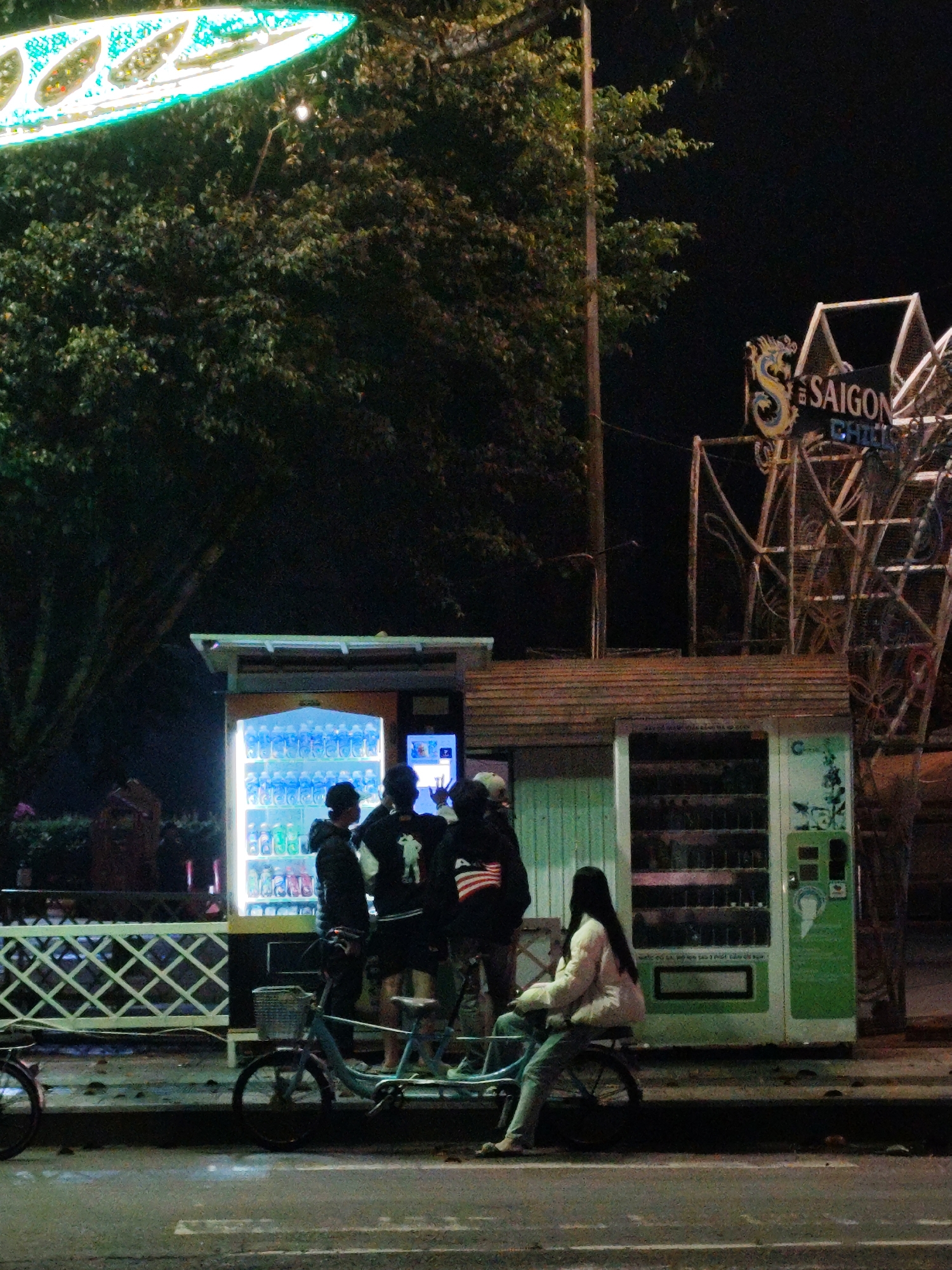 Captured at night in Da Lat, Vietnam, the scene features a group of young people gathered around a brightly lit vending machine. A tandem bicycle sits nearby, occupied by a young woman, seated. Adjacent to the vending machine is a structure with the words “Saigon Chill” displayed above a Ferris wheel-like configuration. Overhead, a brightly lit art piece resembling a kite hangs from a large tree. The street is dark, and a white fence partially obscures the background.