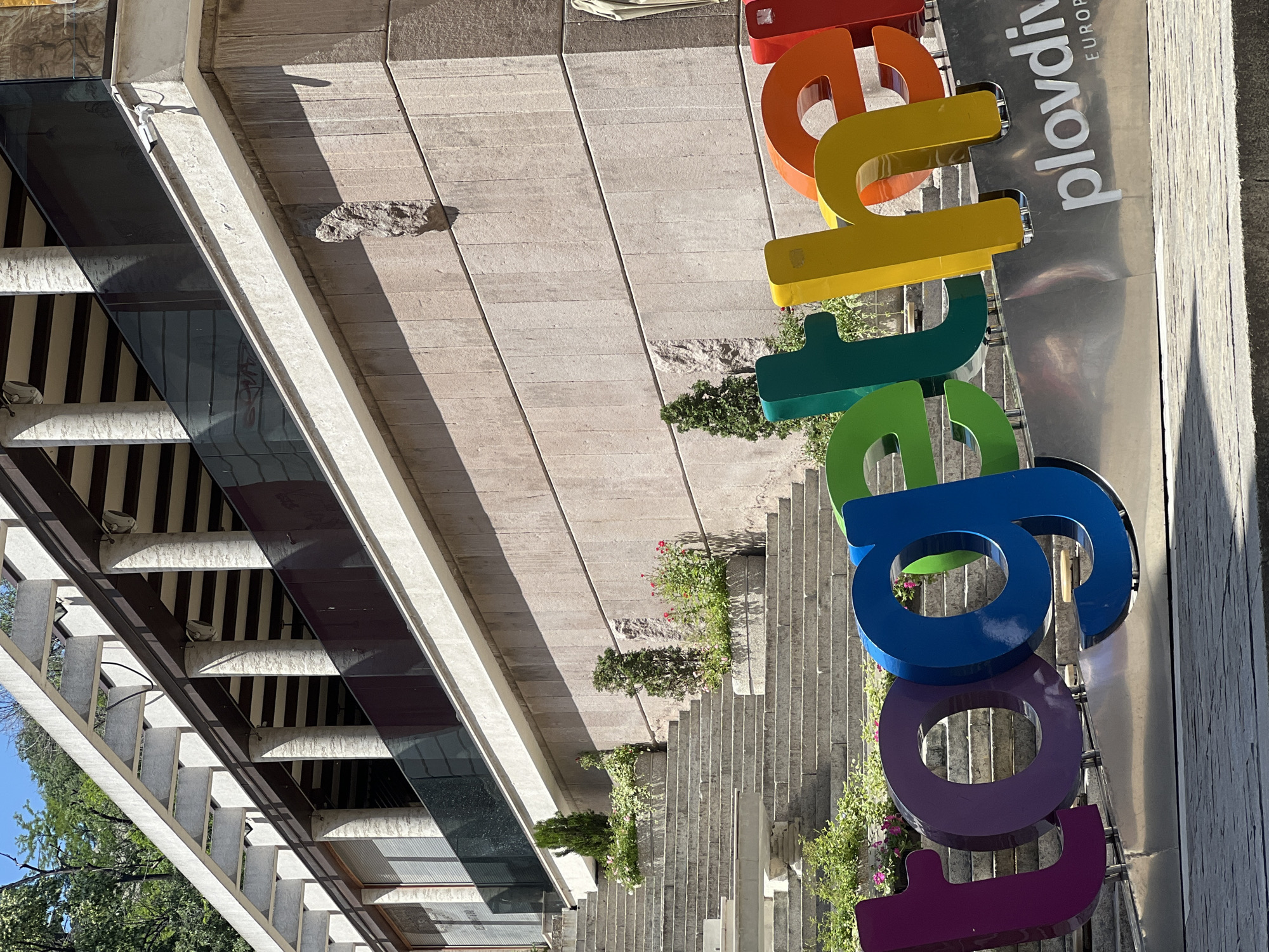 The image captures a street scene in Plovdiv, Bulgaria. It is a ground-level view looking upwards, featuring a modern building on the left. The building's facade includes a glass and concrete structure. Adjacent to this structure is a long staircase, ornamented with greenery and small plants placed on different levels. A large, multi-colored sign, "Plovdiv EUROPEAN," dominates the right side of the frame. The sign's lettering uses various hues and is mounted on a metallic surface. The overall environment is sunny.