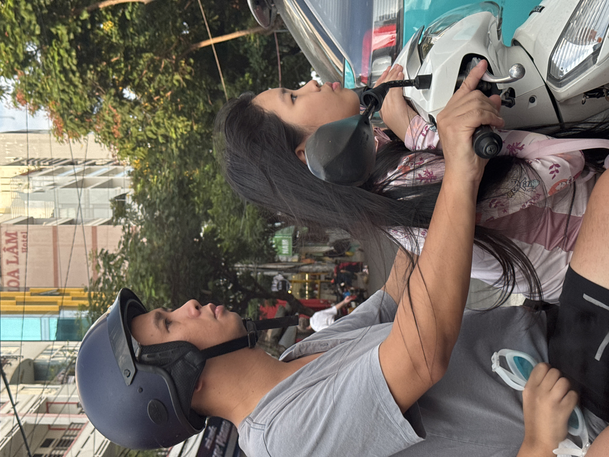 Two individuals, a male and a female, are riding a white motorcycle in Nha Trang, Vietnam. The male is in the foreground, wearing a navy blue helmet and a grey t-shirt. He is holding a pair of white and blue swimming goggles in his left hand. The female is seated behind him, with long black hair, wearing a pink and white patterned top. She is holding onto the handlebars. The motorcycle is white with chrome accents. In the background, buildings line a street, with trees and power lines visible. The buildings appear to be a mix of modern and older architecture, with one prominent building displaying signage in Vietnamese. The lighting suggests it is daytime, possibly late afternoon, with soft sunlight filtering through the trees. The scene appears to be on a city street, likely a busy area given the presence of other vehicles partially visible in the background.