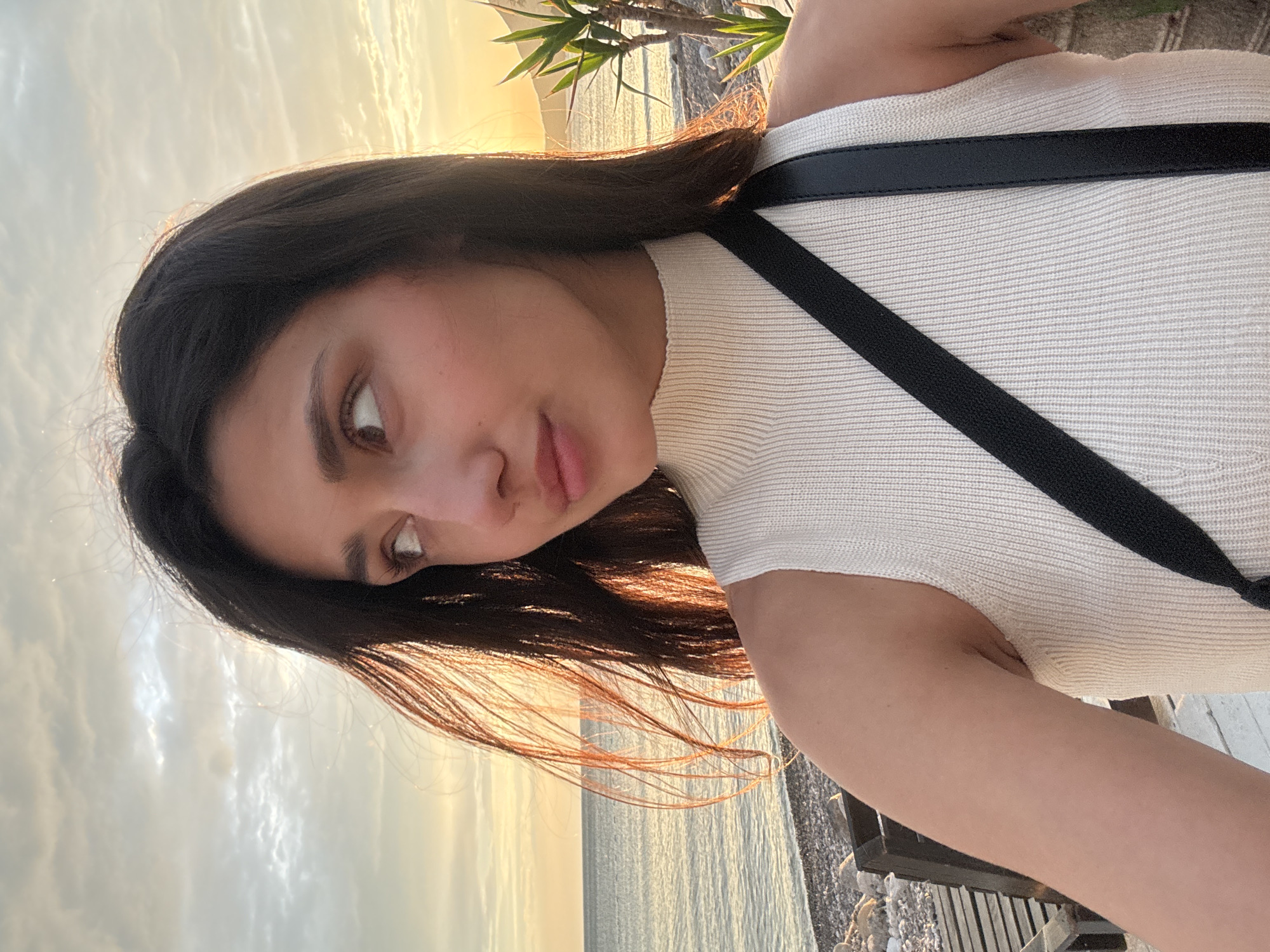 A woman is posing for a selfie. She has dark hair, brown eyes, and fair skin. She is wearing a white ribbed top with a black purse strap. The background shows the sea and sky at sunset, indicating an outdoor location. Based on the beach details, the location is Šušanj, Montenegro.