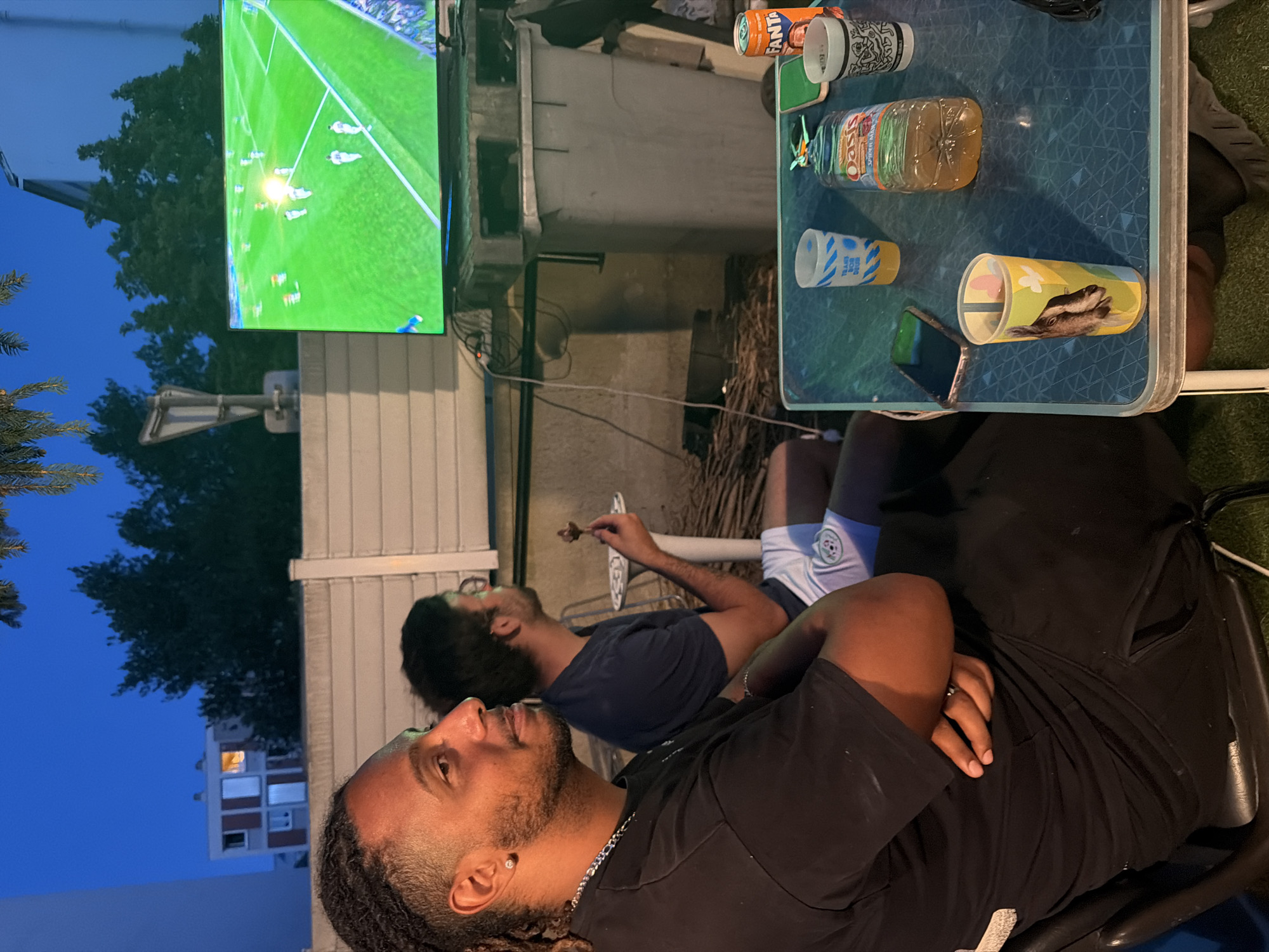 Two men are sitting outdoors at dusk, watching a soccer match on a television screen. The man in the foreground, on the left, is wearing a black t-shirt and has dreadlocks. The man in the background, on the right, is wearing a dark blue t-shirt and white shorts. He is holding a small snack. They are seated at a blue table with drinks and a mobile phone. The setting appears to be a residential area, possibly a backyard or balcony, with buildings and trees visible in the background. The television is placed on top of a large grey bin. The image was taken in Villeurbanne, France, on July 11, 2021.