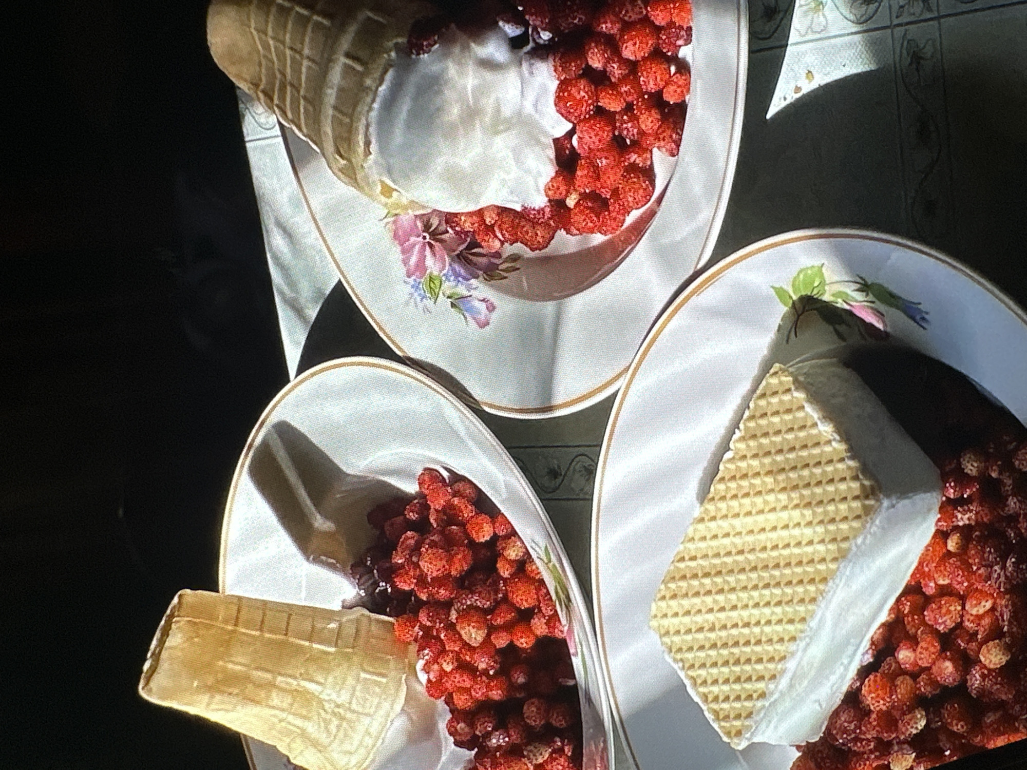 The image shows three white plates with gold trim arranged on a light-colored tablecloth. Each plate contains a dessert. One plate features a waffle cone filled with what appears to be white cream and a pile of small, red berries. Another plate also has a waffle cone alongside a similar pile of berries. The third plate presents a rectangular waffle dessert with a white filling, also surrounded by the same small red berries. The scene is brightly lit, casting shadows across the plates. There is no indication of the location being Yerevan, Armenia. The scene appears to be focused on the desserts and the arrangement of the plates.