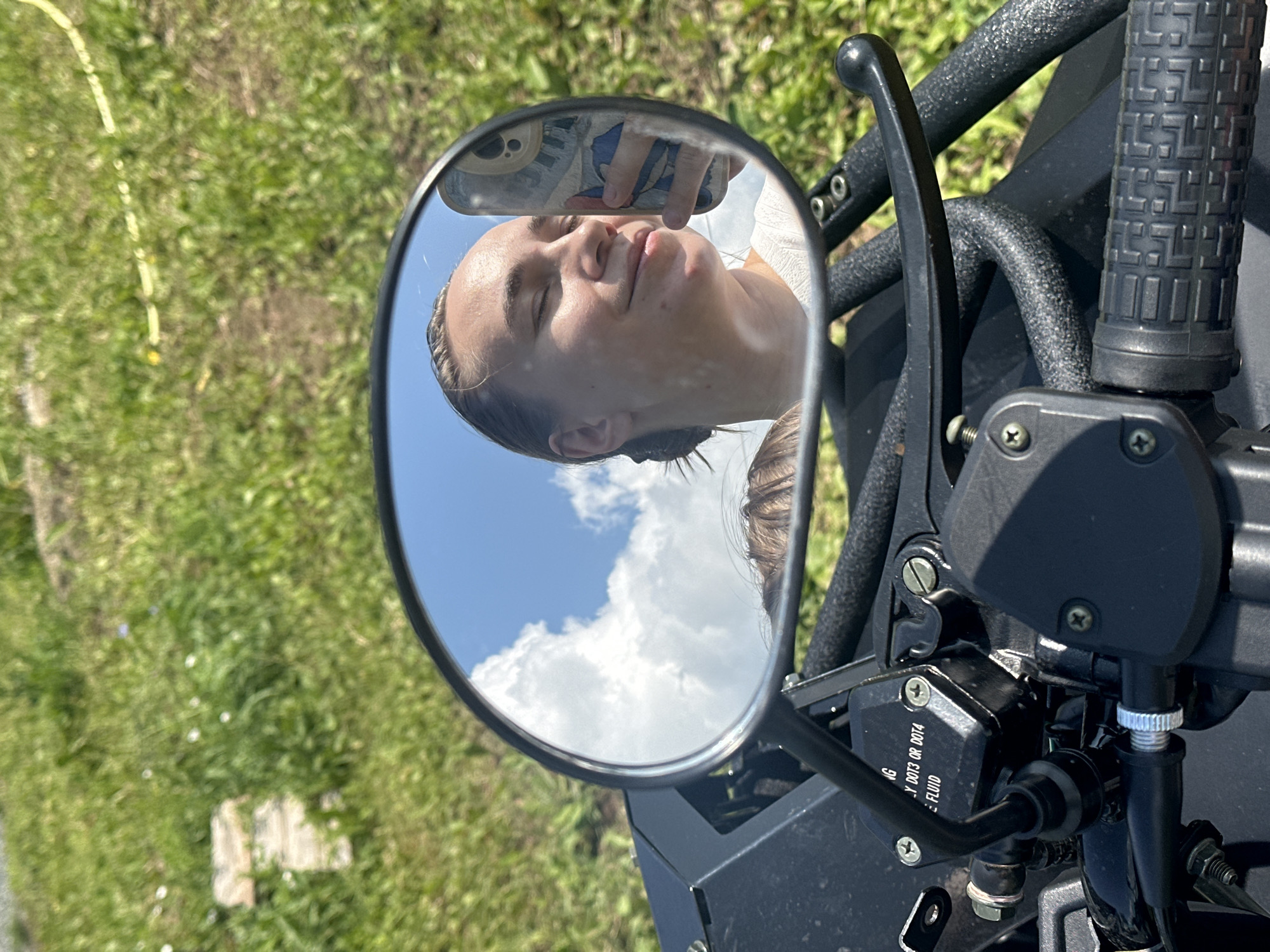 The image shows a close-up, Dutch angle shot of a motorcycle's handlebar and mirror. The mirror reflects a young person holding a phone, taking a selfie. The person has light brown hair pulled back and is looking up with their eyes closed and a slight smile. The reflection also shows a bright blue sky with fluffy white clouds.
The motorcycle's handlebar is black with a textured grip and a black lever. The mirror is round with a black frame. In the foreground, part of the motorcycle's bodywork is visible, painted black. The background is a blur of green grass and some yellow material, possibly a hose or wire.
The lighting suggests it's daytime, likely sunny due to the clear blue sky in the reflection. The scene appears to be outdoors, in a grassy area. There is some text visible on a black component near the handlebar: "NG Y DOT3 OR DOT4 E: FLUID". This indicates it's a brake fluid reservoir, likely for a vehicle. The location context is Ольховка, Russia. The overall mood conveyed is relaxed and casual, with the person enjoying a sunny day.
